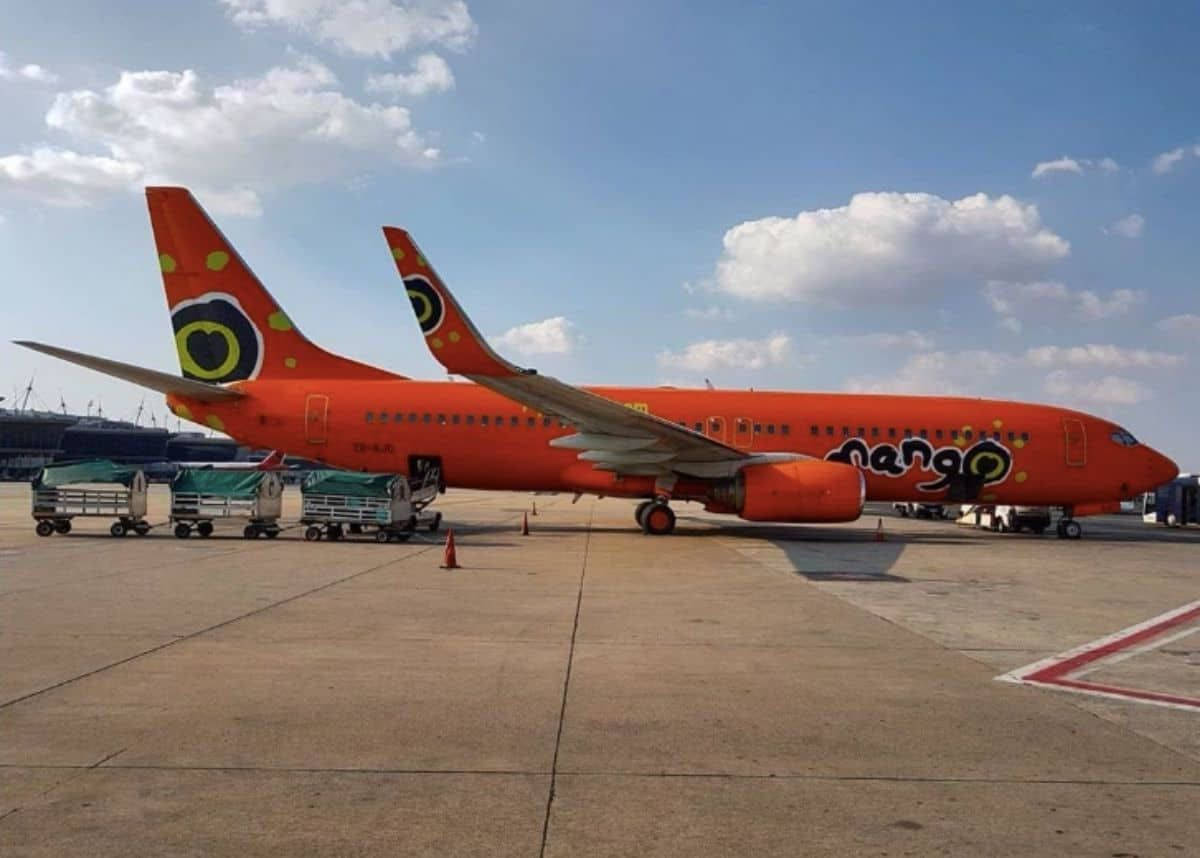 橙色芒果航空飛機停在機場停機坪上，有貨運手推車和藍天。