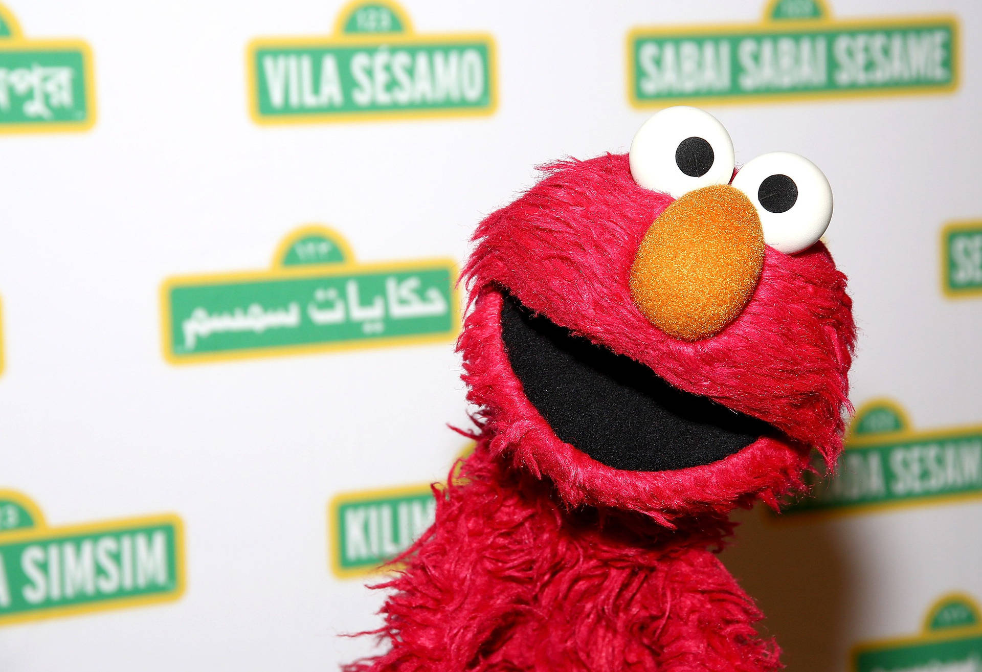 紅色毛絨絨的 Elmo 木偶微笑，背後有芝麻街標誌。