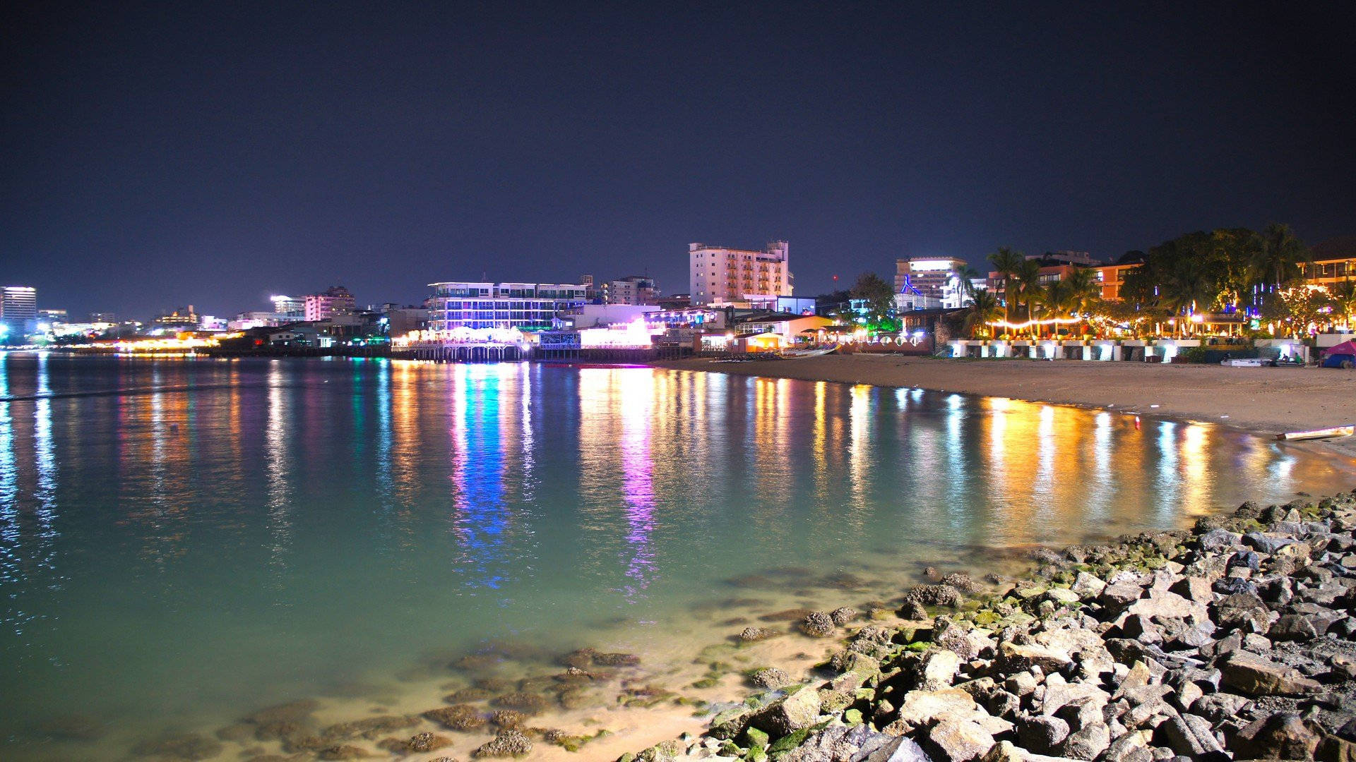 芭堤雅夜間岩石海灘 壁紙 — 夜間海岸城市景觀，鮮豔的霓虹燈光映照在岩石海岸旁的平靜水面上。