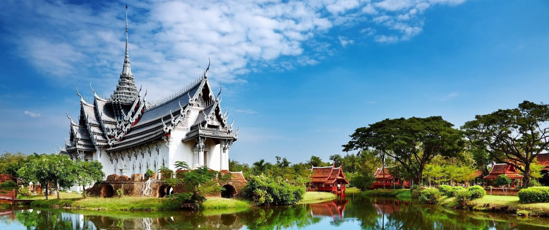 芭堤雅寺廟 壁紙 — 一座宏偉的白色泰國寺廟，帶有華麗的尖頂，在晴朗藍天和蓬鬆白雲下，倒映在寧靜的池塘中。