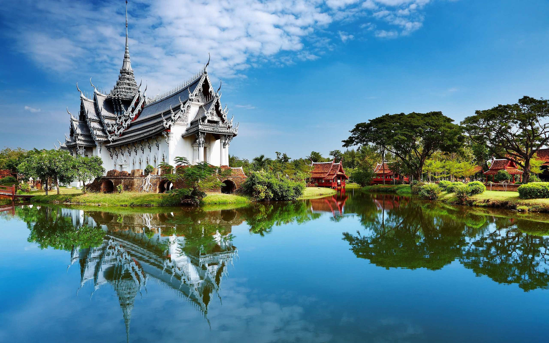 芭堤雅寺廟湖面倒影 壁紙 — 一座令人驚嘆的泰國寺廟，擁有精緻的尖頂，在明亮的天空下完美地倒映在平靜的藍色池塘中。