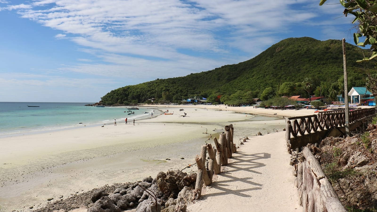 芭堤雅海灘沙岸 壁紙 — 陽光明媚的熱帶海灘，有白色沙灘、碧綠海水和點綴著色彩繽紛建築物的茂密綠色山坡。