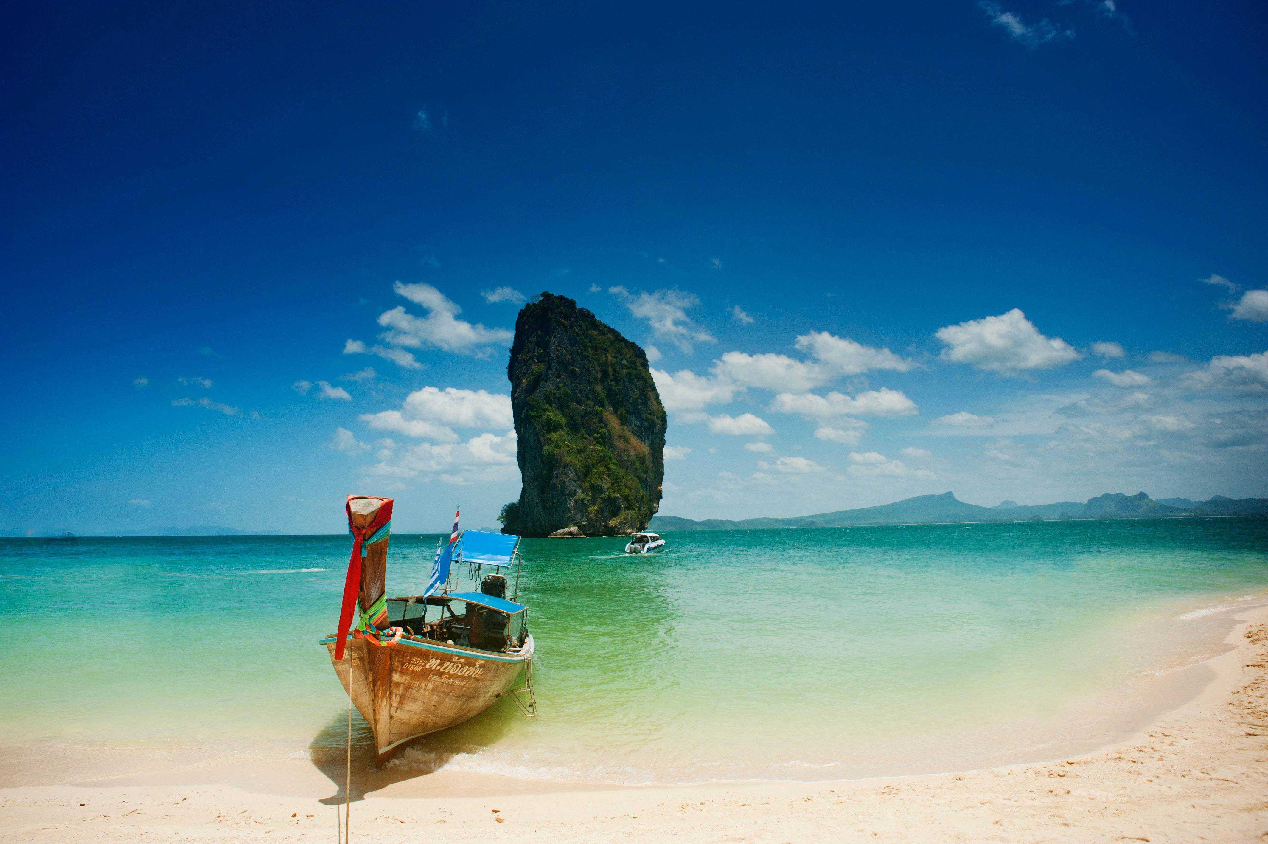 芭堤雅海灘泊船的風景 壁紙 — 一艘傳統長尾船停泊在 pristine 白色沙灘上，碧綠海水和遠處高聳的石灰岩喀斯特地貌在明亮的藍天下。