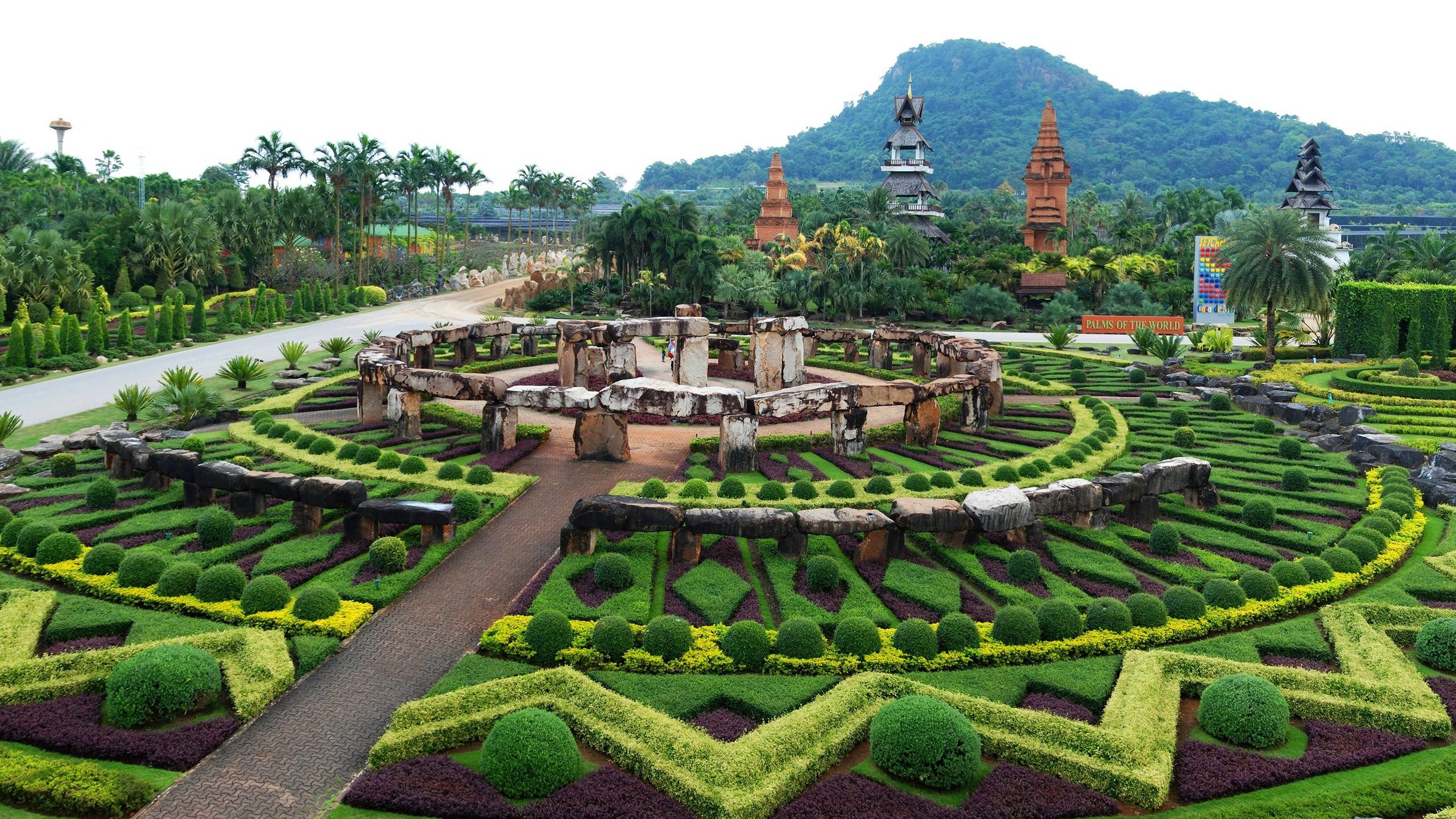 芭堤雅熱帶海灘風景 壁紙 — 從空中俯瞰一個精心修剪的花園，幾何形狀的樹籬和石頭廢墟被茂密的熱帶植物和遠處的寺廟式建築所環繞。
