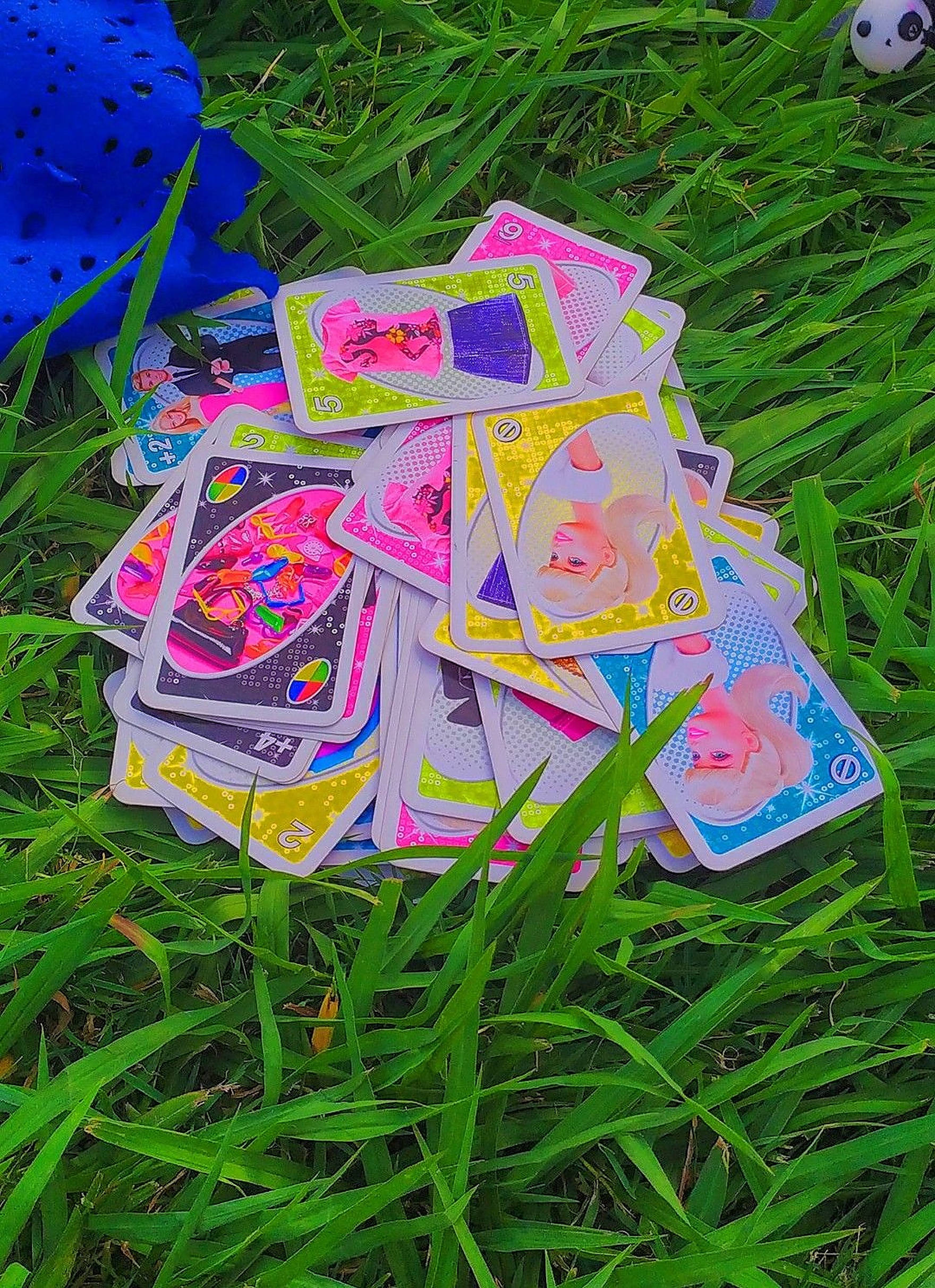 芭比 Uno 卡牌 壁紙 — 色彩繽紛的卡通卡片散落在茂盛的綠色草地上，附近有一個藍色毛絨玩具。