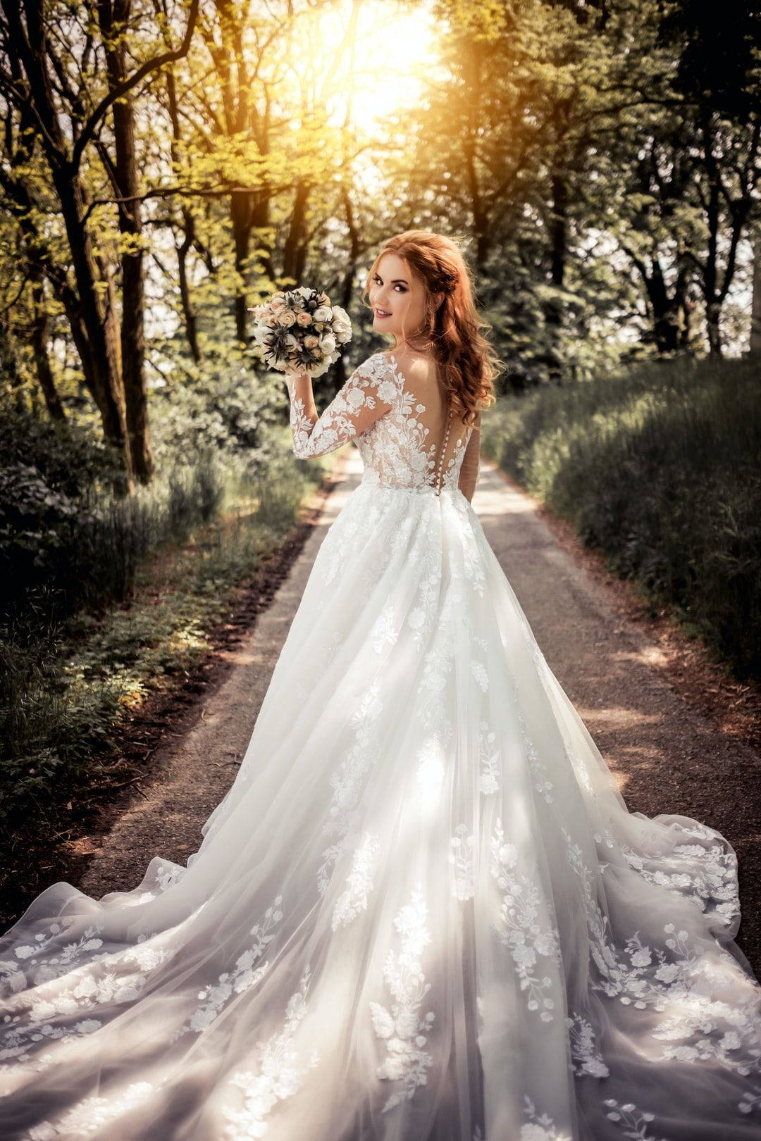 花園中的迷人新娘 壁紙 — 一位身穿白色蕾絲婚紗的新娘，手持花束，走在陽光明媚的森林小徑上。