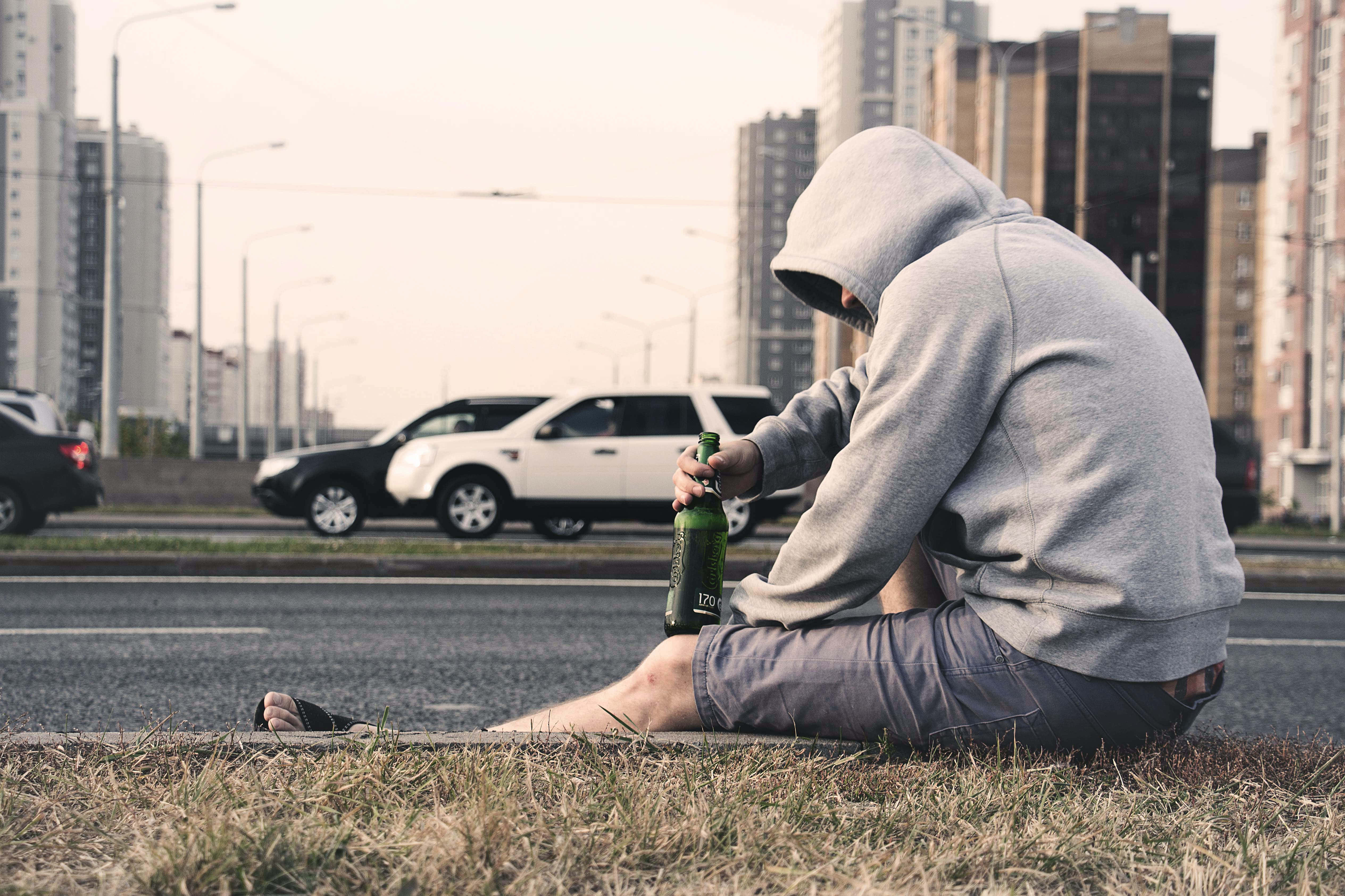 「苦難中的男子：永恆痛苦的肖像」 壁紙 — 一個戴連帽衫的人獨自坐在路邊，手持啤酒瓶，汽車在都市景觀中駛過。