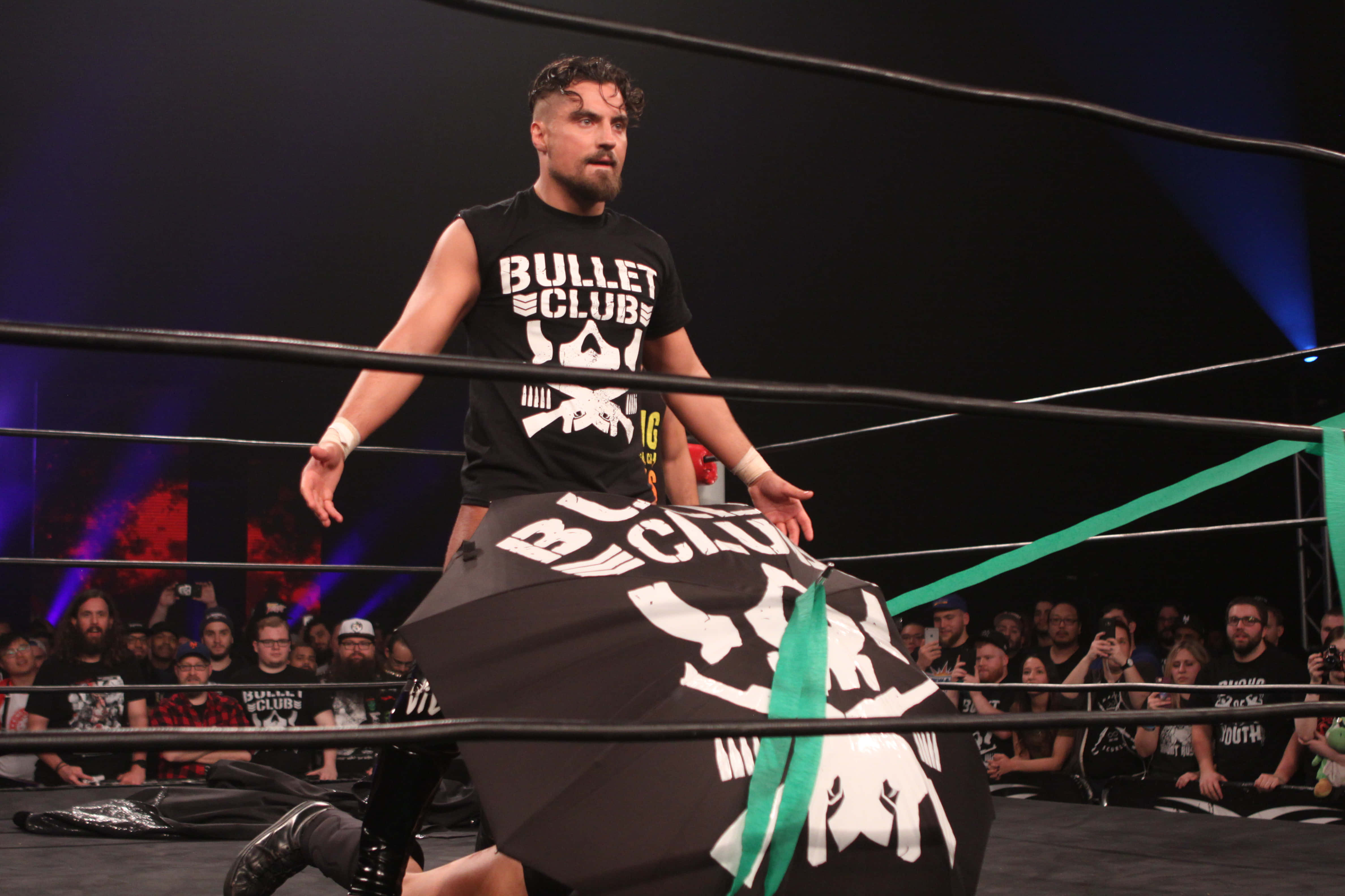 英國ROH摔角手馬蒂·斯卡爾爾與亞當·柯爾 壁紙 — 一名身穿黑色 'Bullet Club' T恤的摔角手站在擂台上，手持相配的旗幟，粉絲們在舞台燈光下觀看。