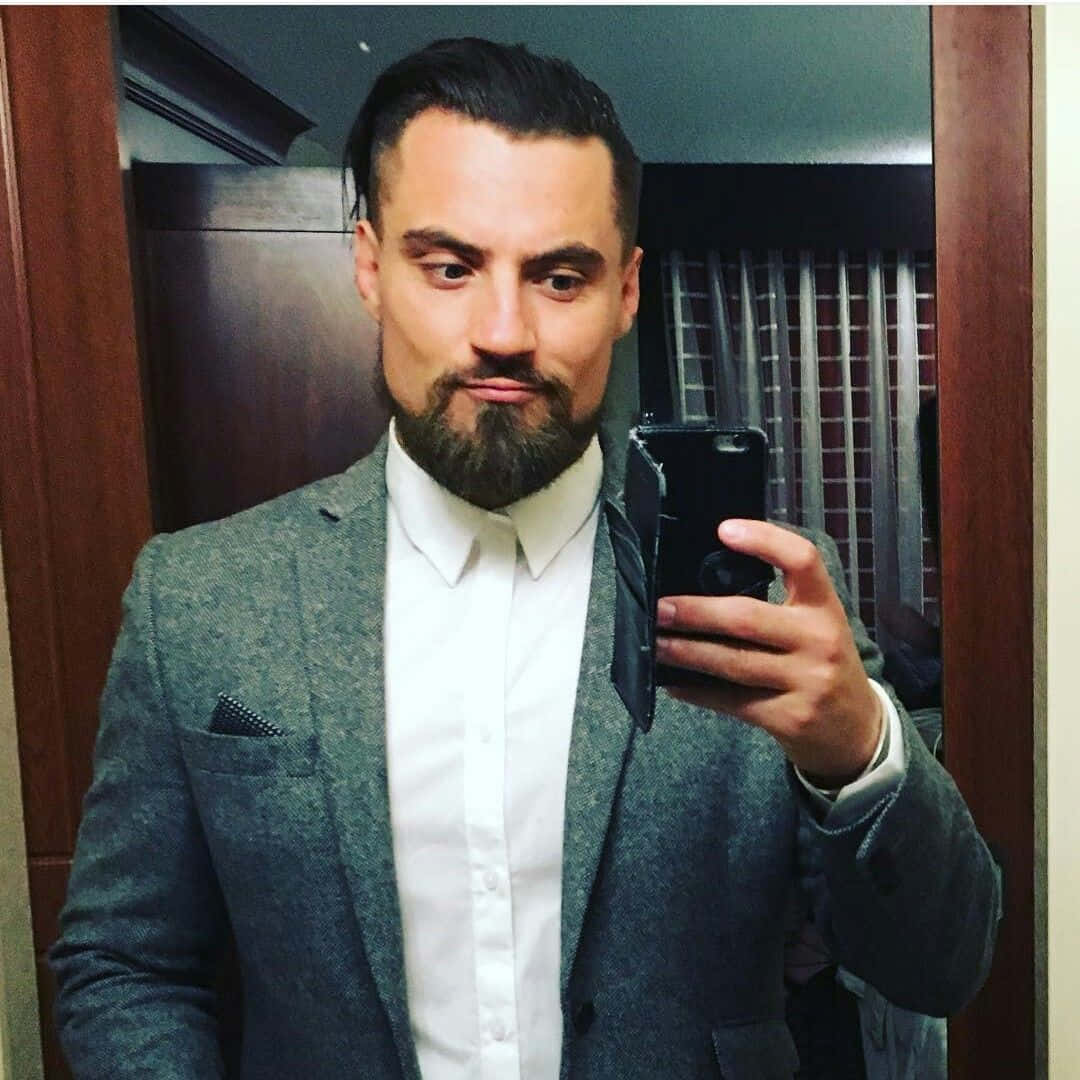 英國ROH摔角手馬蒂·斯卡爾鏡前自拍 壁紙 — 一位留鬍鬚的男人在室內穿著灰色西裝和白色襯衫對著鏡子自拍。