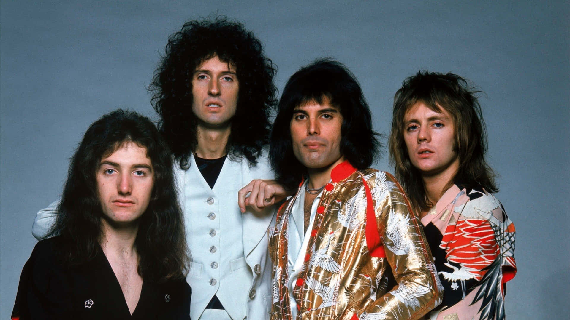 英國傳奇搖滾樂隊Queen自1970年代起震撼世界 壁紙 — 四位標誌性搖滾音樂家穿著華麗的70年代服裝，在樸素的灰色工作室背景前自信地擺姿勢。