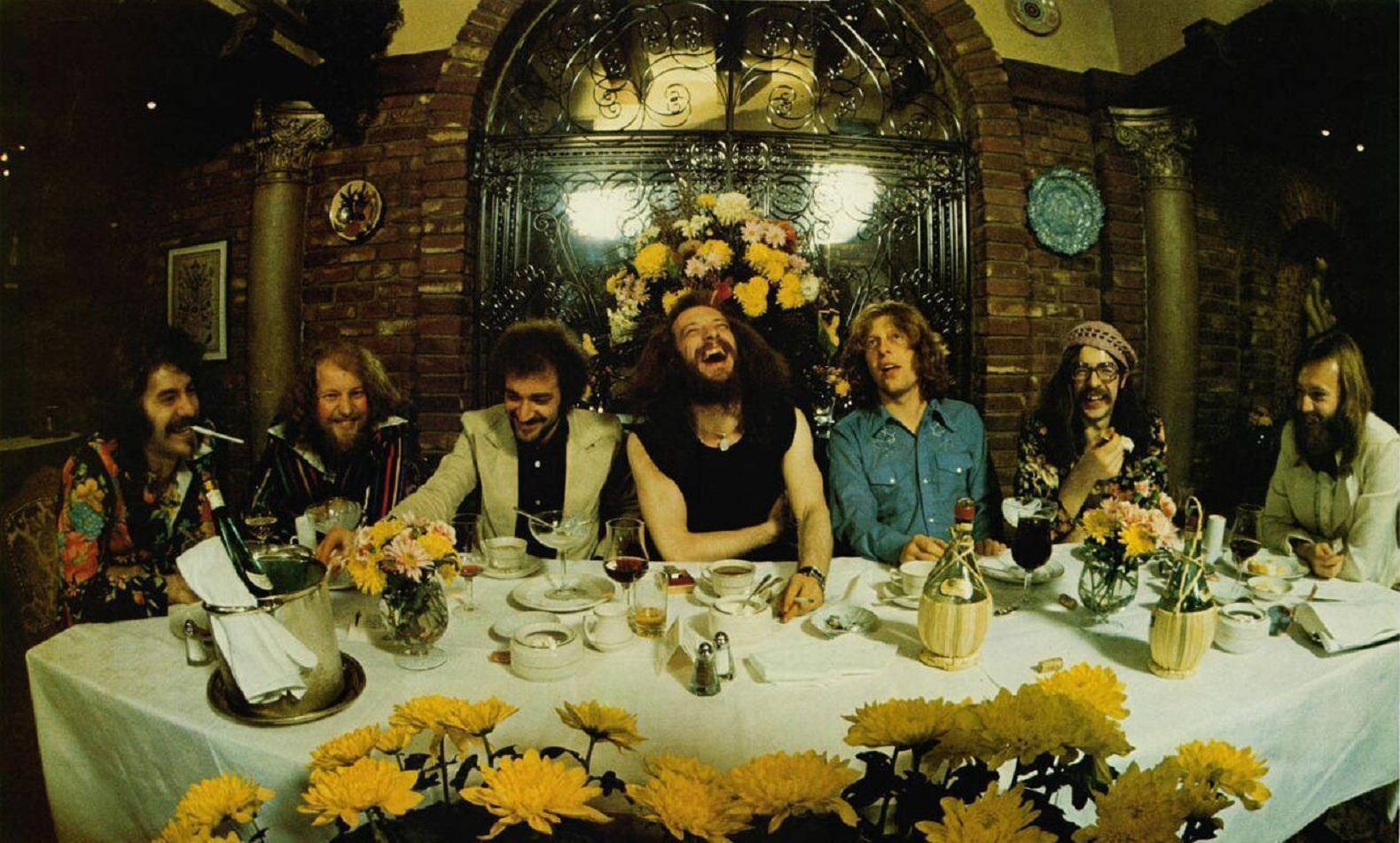 英國前衛搖滾樂隊 Jethro Tull 古風魚眼鏡頭拍攝 壁紙 — 一個歡樂的樂隊在擺滿黃色花朵和復古裝飾的豐盛餐桌旁一起大笑。