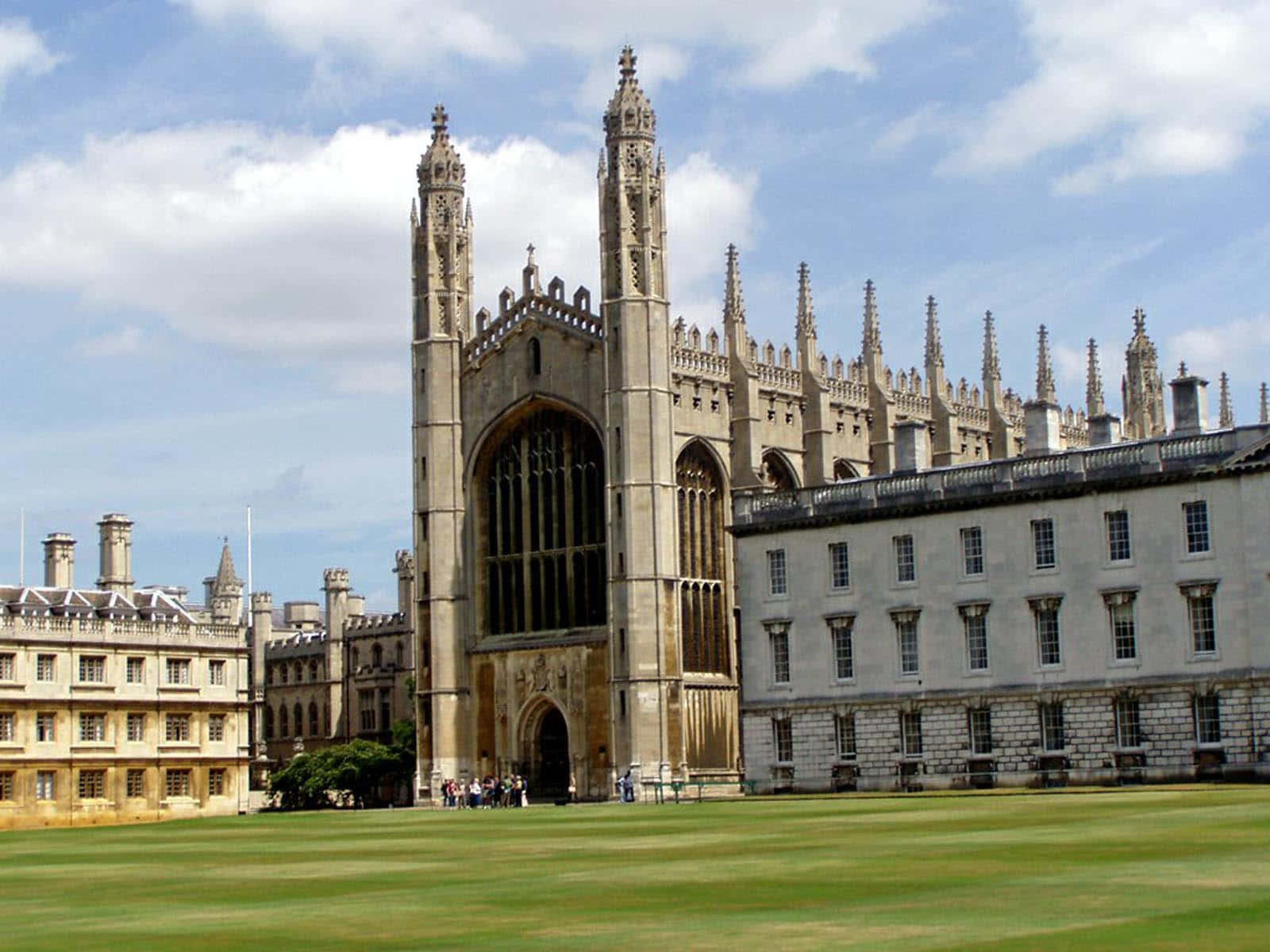 英國劍橋大學王后學院莊嚴風貌 壁紙 — 宏偉的哥德式教堂，雙塔高聳入雲，襯著藍天，兩旁是歷史悠久的石造建築和修剪整齊的綠色草坪。