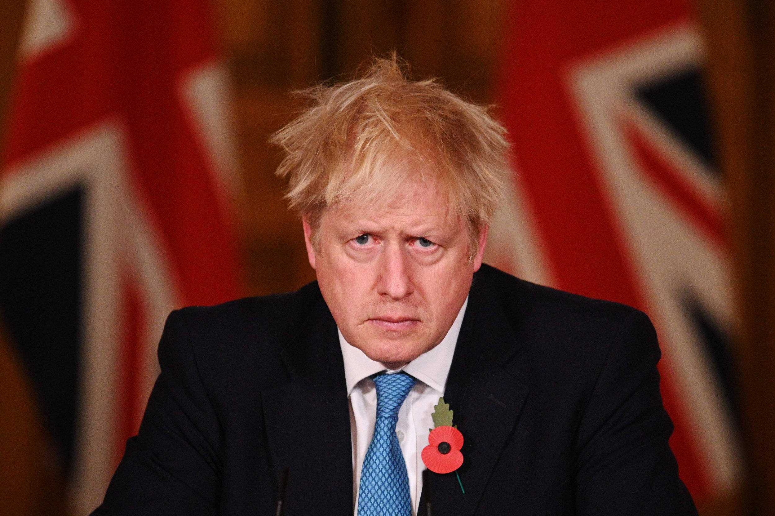 英國國旗後的 Boris Johnson 壁紙 — 一名身穿深色西裝和藍色領帶的男子，在模糊的英國國旗背景下佩戴紅色罌粟花胸針。
