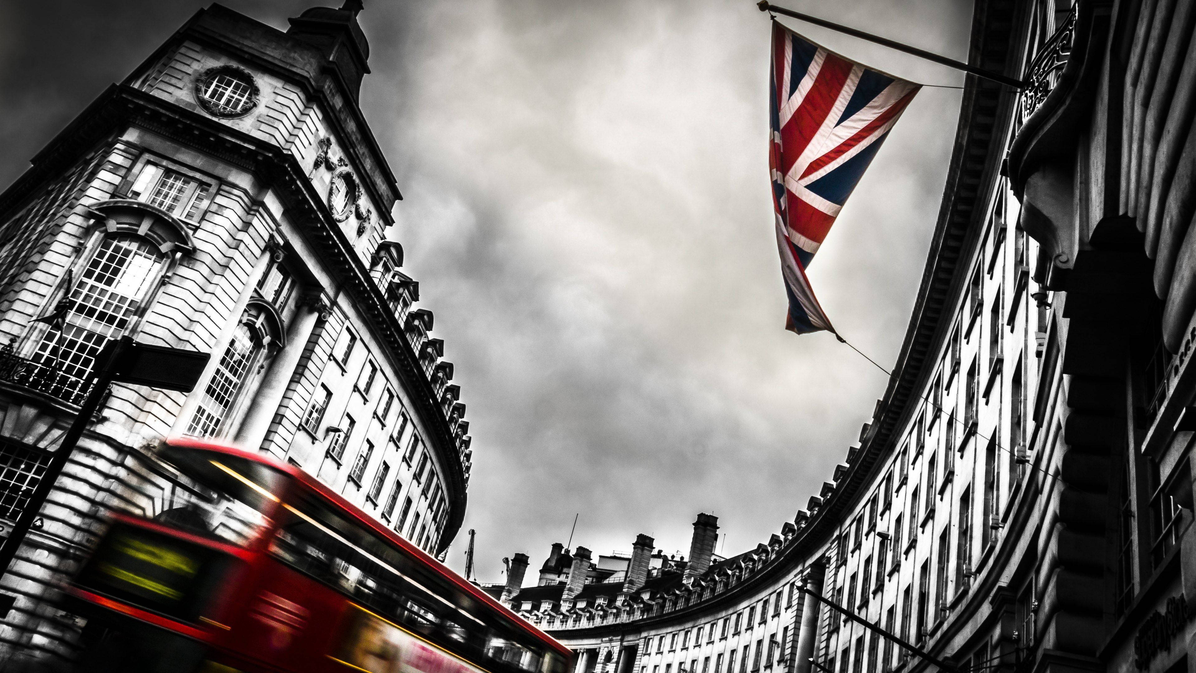英國國旗黑白經典圖案 壁紙 — 低角度拍攝一輛紅色雙層巴士在陰沉天空下，帶有飄揚的英國國旗，飛馳經過歷史悠久的倫敦建築。