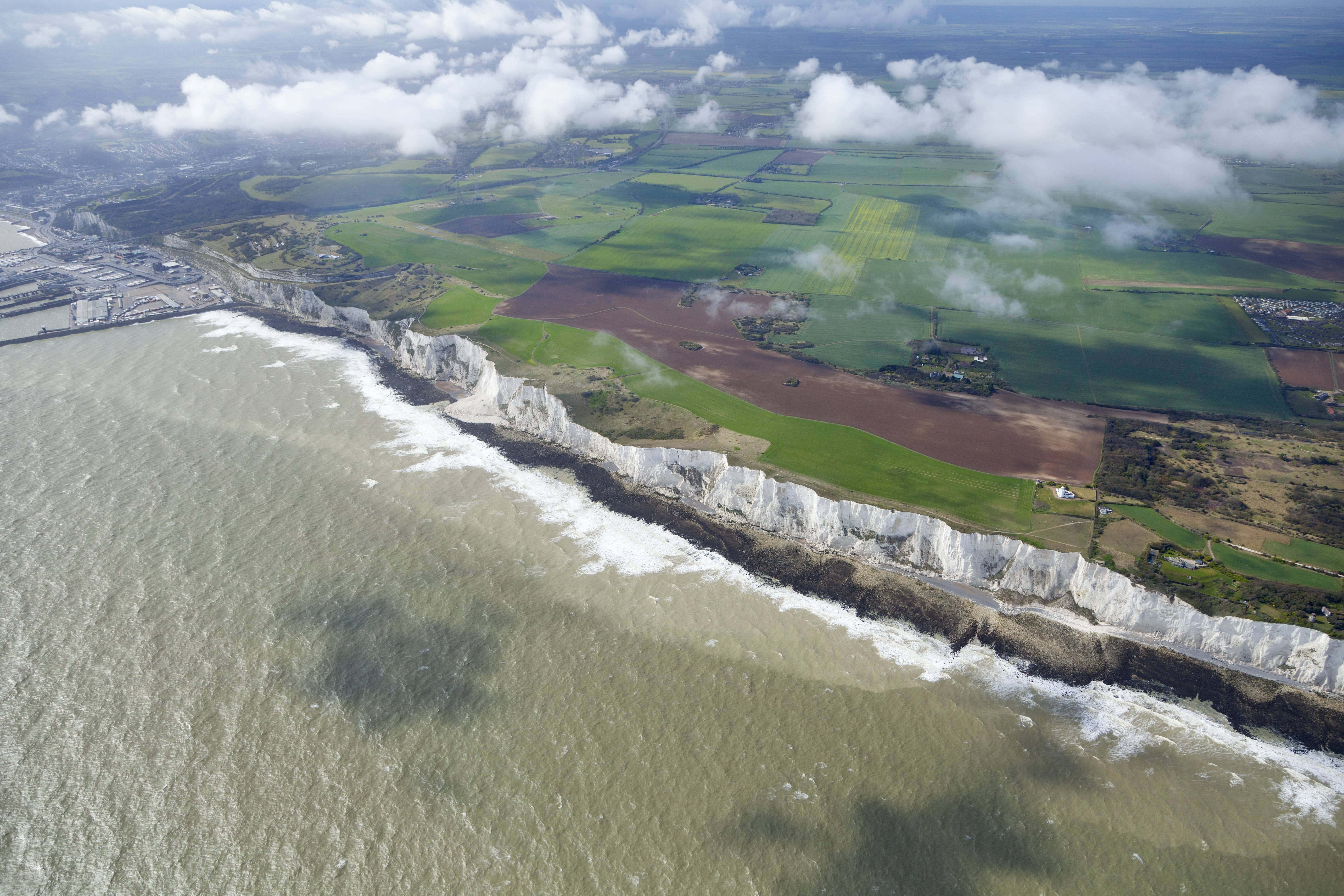 英國多佛白崖空中拍攝 壁紙 — 戲劇性的白色白堊懸崖與洶湧的灰色海洋交匯的鳥瞰圖，上方有拼布般的綠色田野和散落的雲朵。
