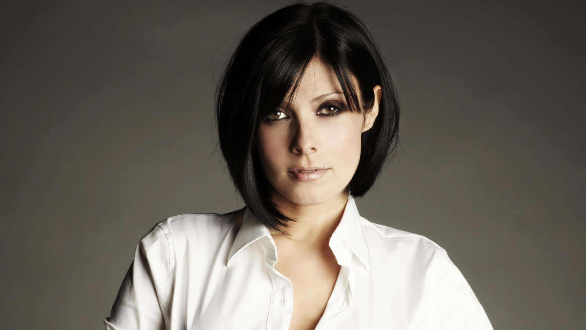 英國女演員Kym Marsh 壁紙 — 一位短黑髮、煙燻妝眼睛的女性，穿著一件清爽的白色鈕扣襯衫，在柔和的灰色背景前直視鏡頭。