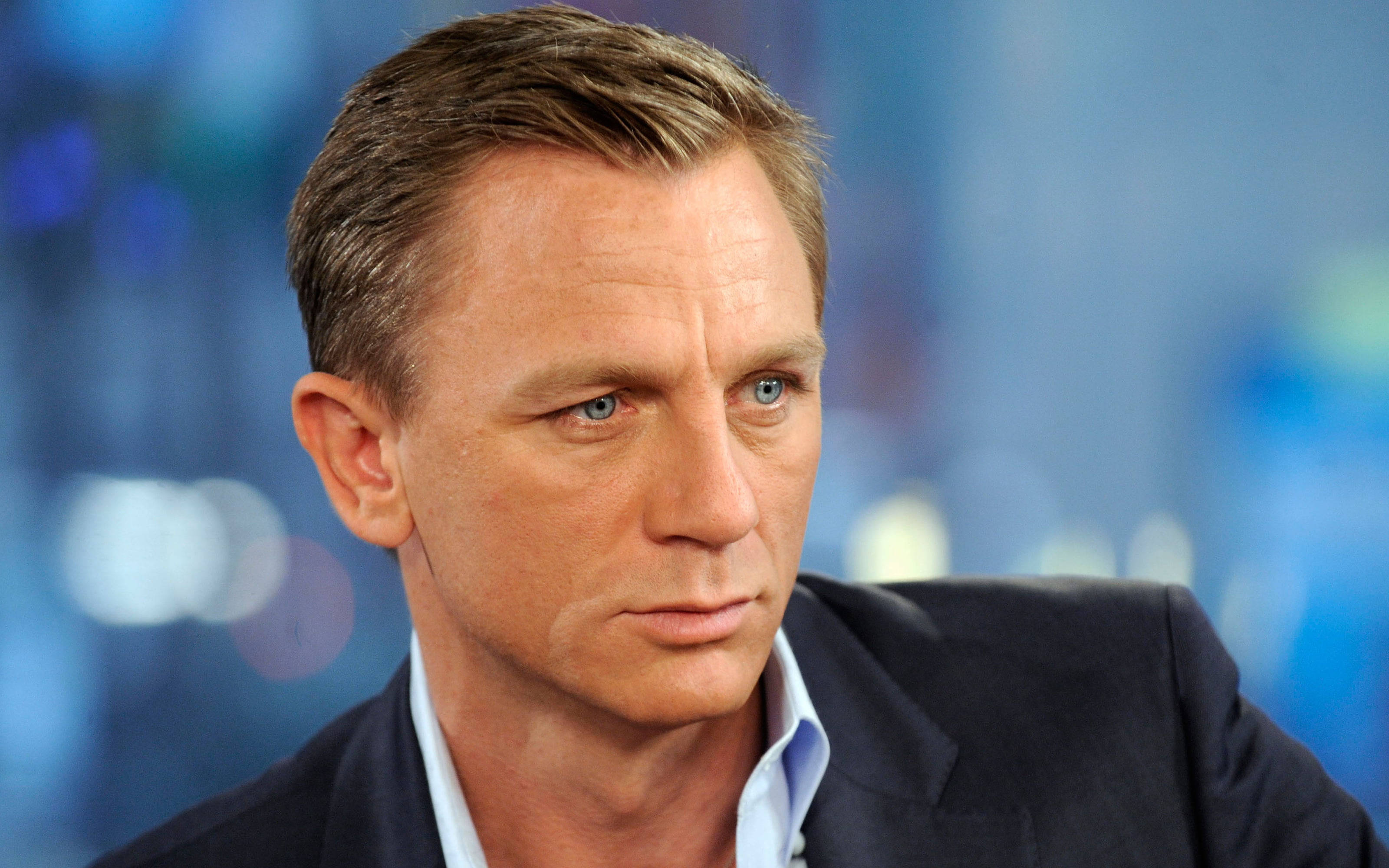 英國明星丹尼爾·克雷格 壁紙 — 丹尼爾·基克（Daniel Craig）的頭像特寫，擁有強烈的藍色眼睛和嚴肅的表情，背景為模糊的藍色。