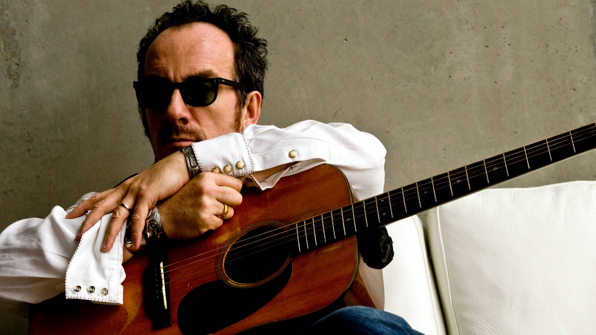 英國格萊美得主Elvis Costello 壁紙 — 一名戴太陽眼鏡的男子坐在白色梳化上，頭靠著木結他，背景是混凝土牆。