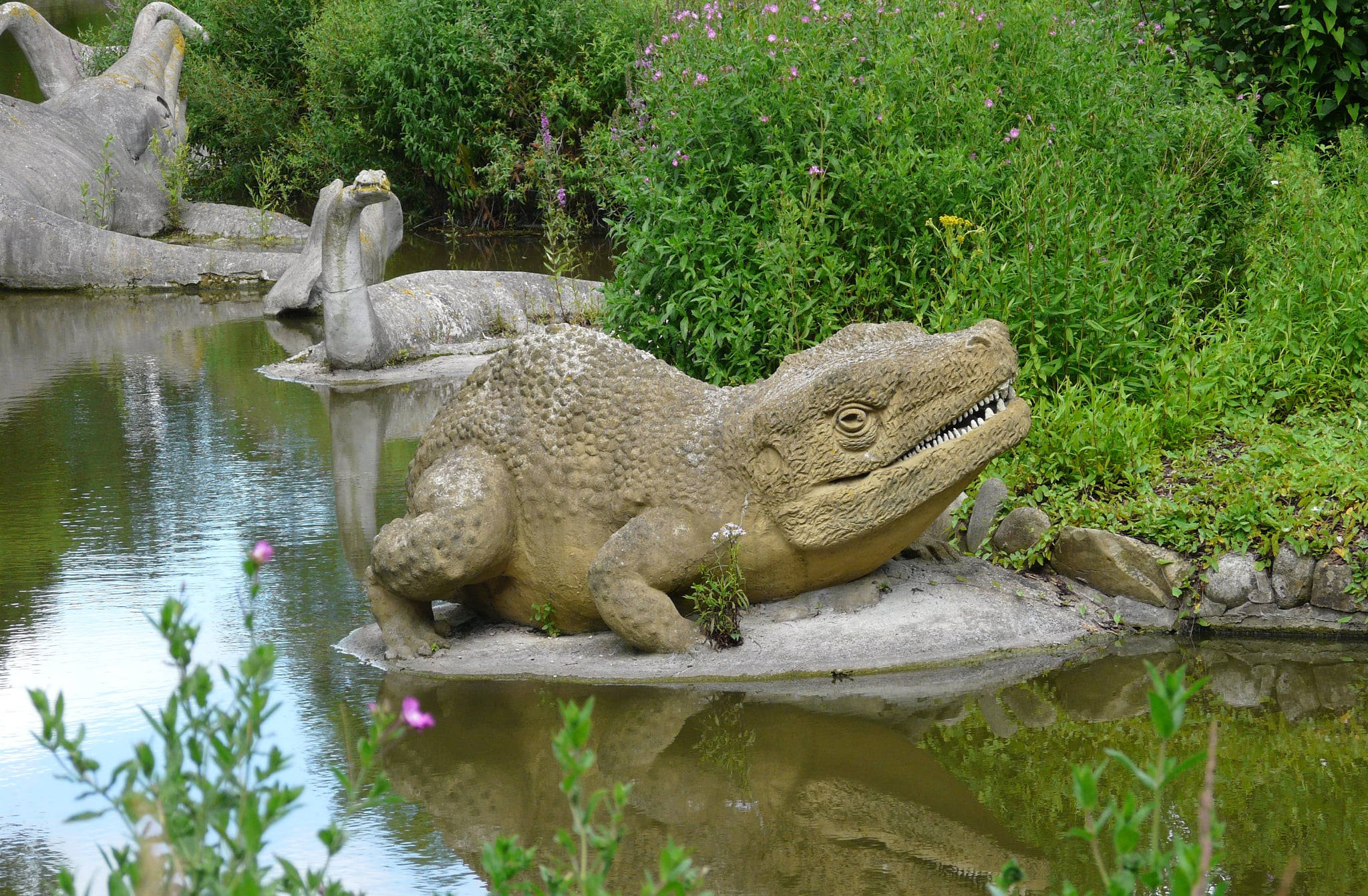 英國水晶宮花園 壁紙 — 一個大型石鱷魚雕像放置在池塘邊，周圍環繞著茂密的綠色植物和紫色花朵。