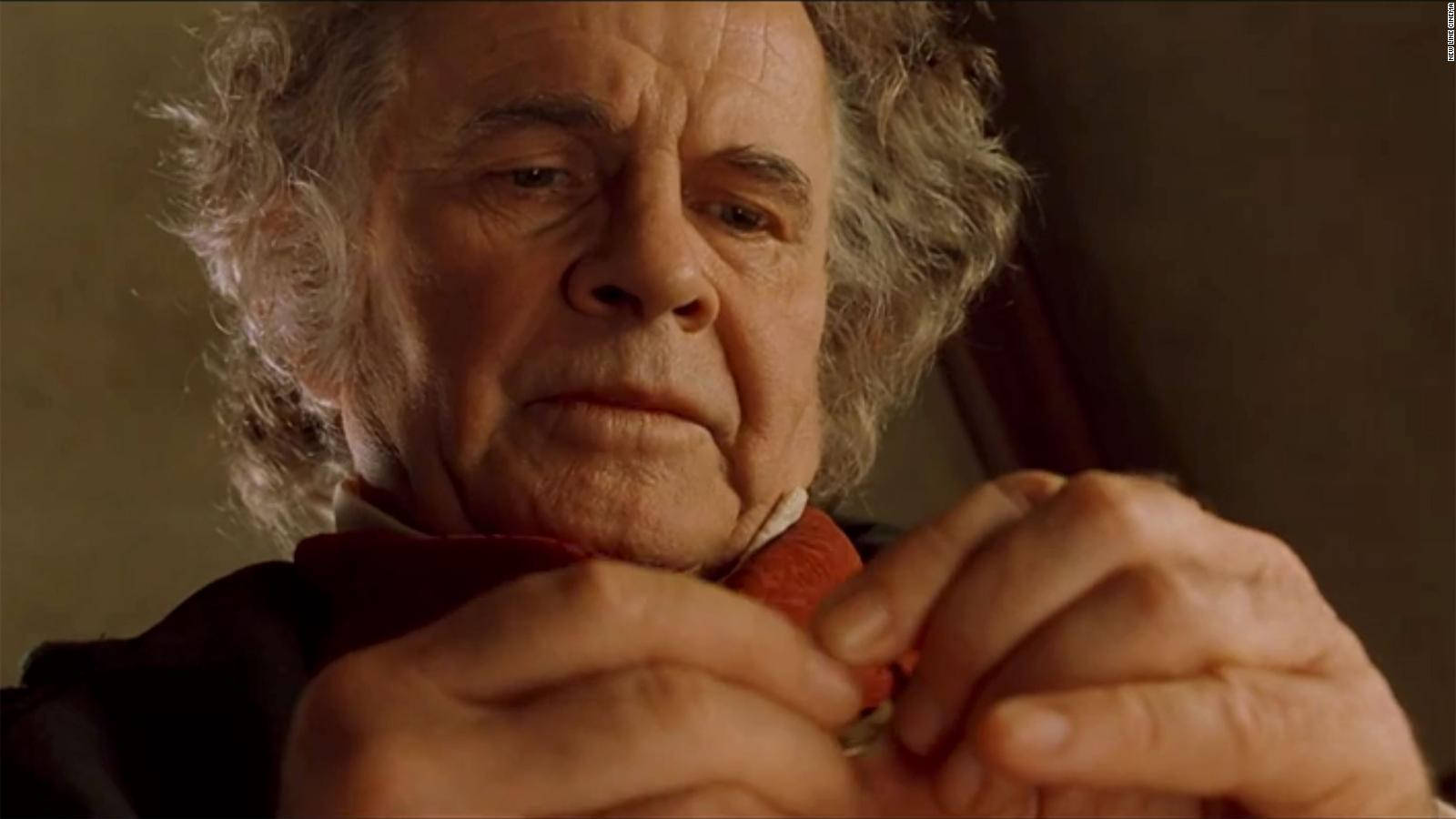 英國演員伊恩·霍姆在《魔戒： fellowship of the Ring》中飾演比爾博·巴金斯 壁紙 — 一位灰白鬈髮的長者，在溫暖燈光下專注地檢查手中的小物件。