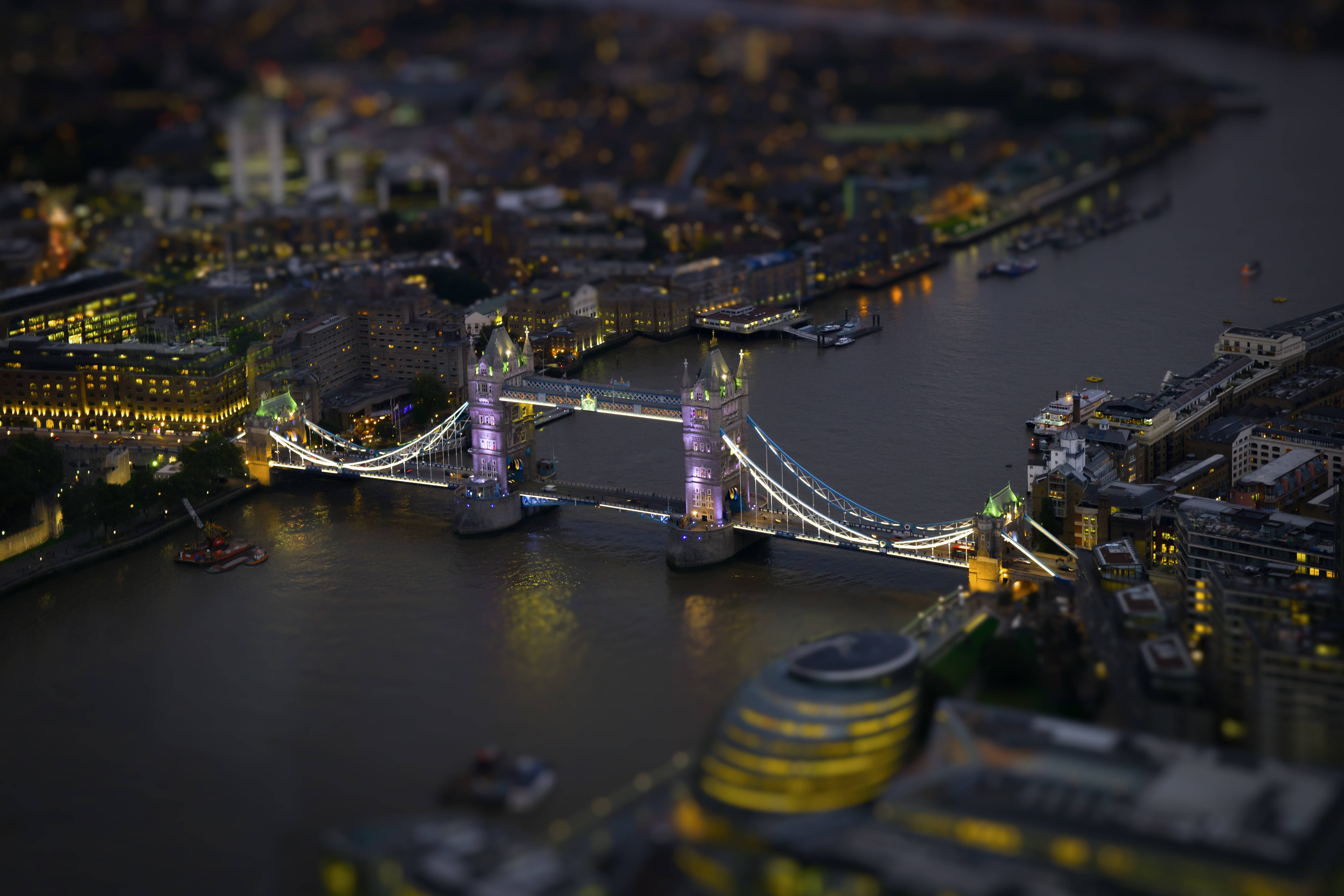 英國無人機拍攝塔橋 壁紙 — 倫敦塔橋在黃昏時的微縮移軸視角，發光的燈光倒映在河面上。