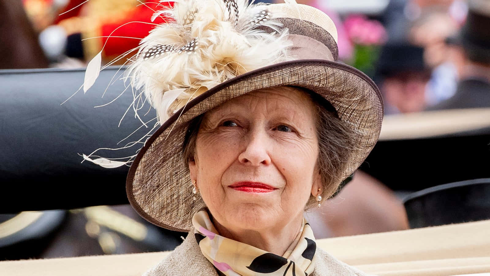 英國皇室安妮公主出席年度阿斯科特賽馬場 壁紙 — 一位優雅的老年女士戴著一頂飾有羽毛和圖案圍巾的寬邊草帽，坐在戶外。