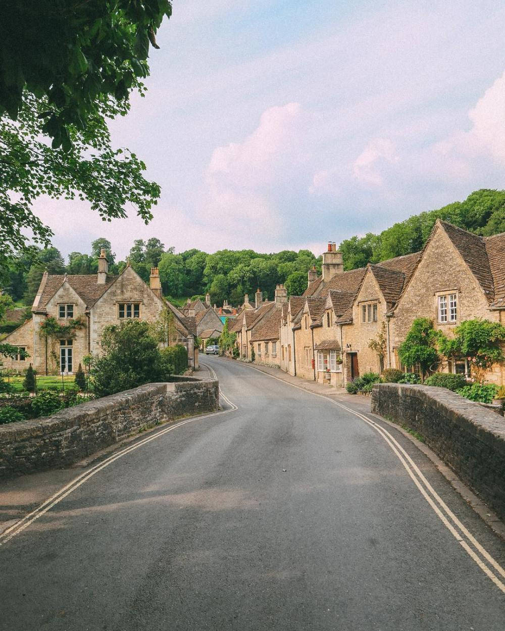 英國科茨沃爾茲地區蜿蜒道路 壁紙 — 一條蜿蜒的道路穿過一個迷人的村莊，村莊裡有石頭小屋和茂密的綠樹，天空多雲。