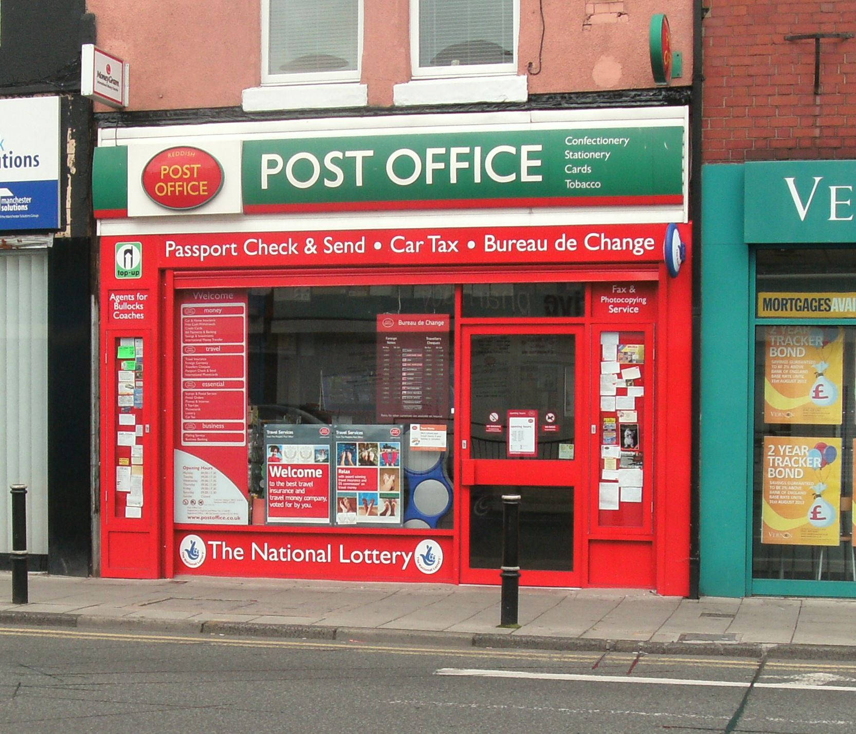 英國雷迪什郵政局 壁紙 — 充滿活力的紅色郵政局店面，玻璃窗展示服務和彩票廣告，位於城市街道上。