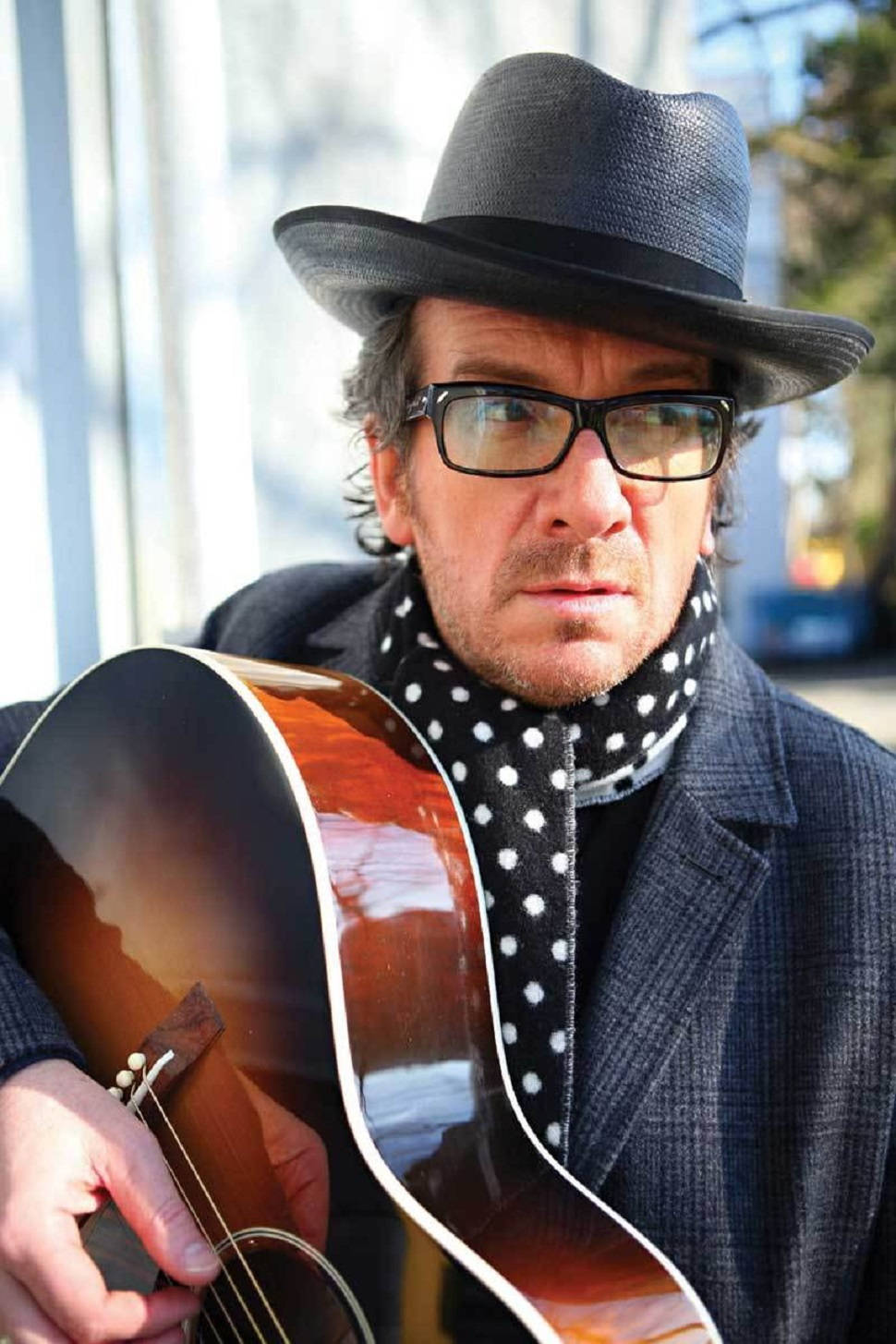 英國音樂人Elvis Costello 壁紙 — 一個戴黑色紳士帽和眼鏡的男人，在明亮的窗戶背景下拿著一把陽光照射的木結他。