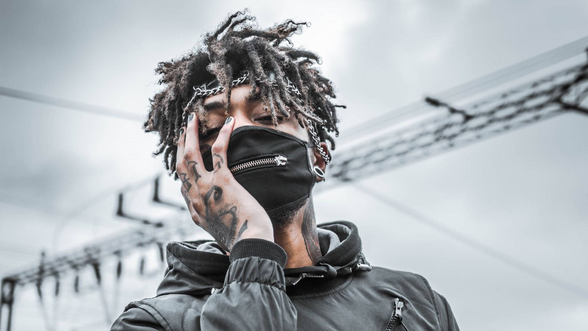 英國饒舌歌手 Scarlxrd 熱情演出 壁紙 — 一個有辮子頭和紋身的人用黑色口罩遮住臉，背景是灰色天空和電線。