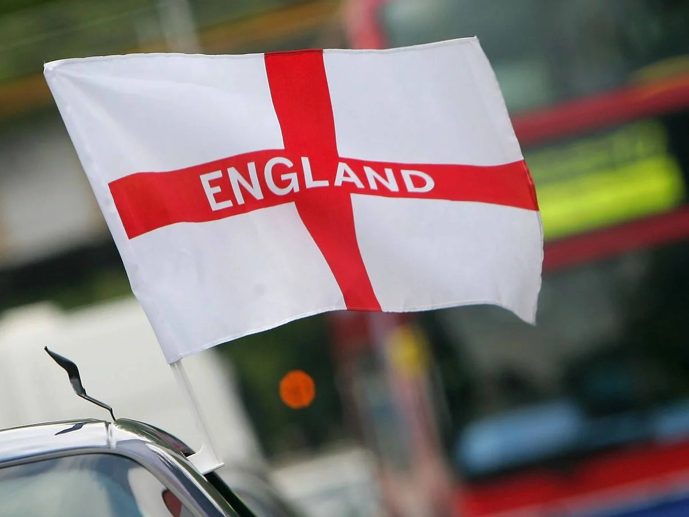 英格蘭國旗聚焦特寫 壁紙 — 一面帶有 'ENGLAND' 字樣的紅色十字旗在汽車天線上飄揚，背景是模糊的城市景觀。