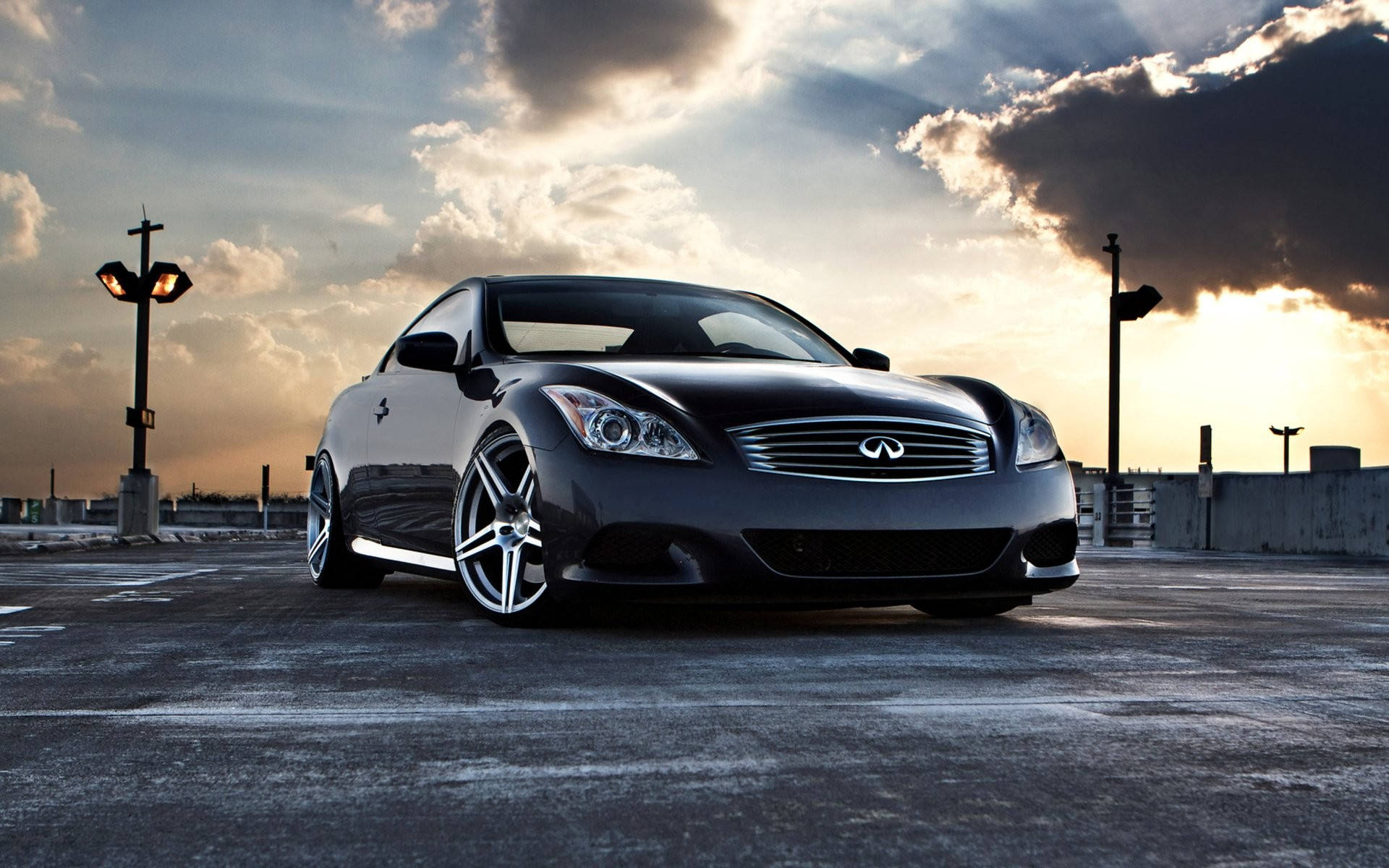 英菲尼迪 G37 車型 壁紙 — 一輛時尚的黑色Infiniti雙門跑車停泊在日落時分的屋頂上，背景有戲劇性的雲彩和發光的街燈。