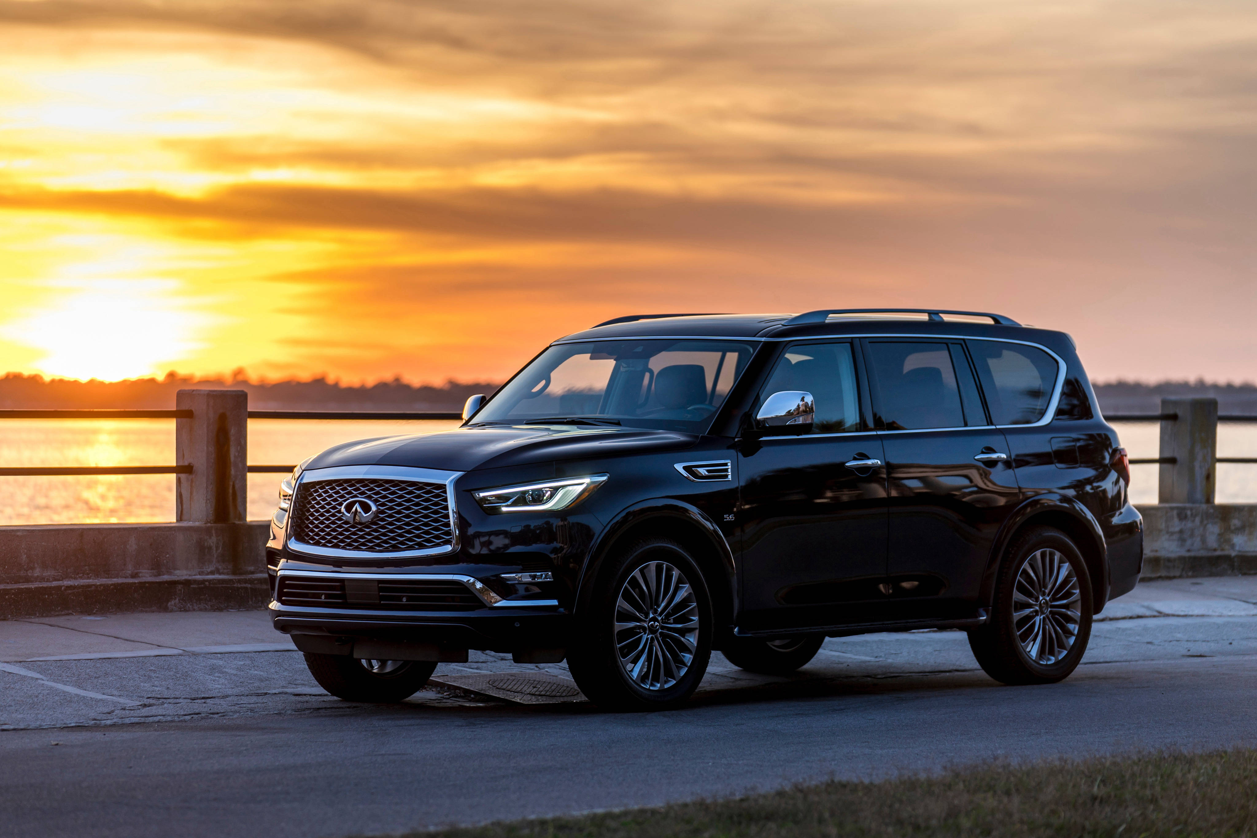 英菲尼迪SUV與日落 壁紙 — 一架時尚黑色 Infiniti QX80 SUV 停泊在海濱碼頭，背景是金色夕陽和溫暖的橙色雲彩。