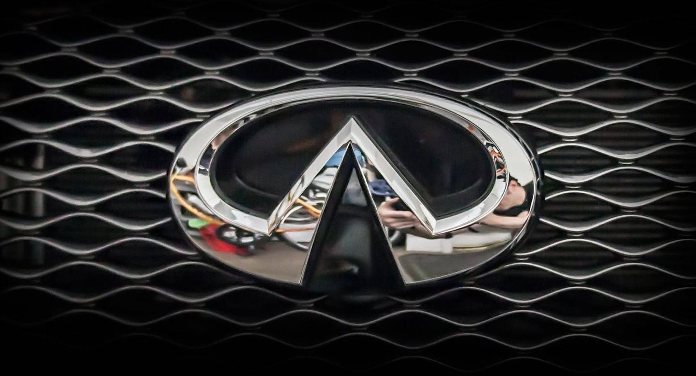 英菲尼迪車標特寫 壁紙 — 閃亮鍍鉻Infiniti標誌居中於黑色波浪形格柵上，反射表面顯示模糊的周圍環境。