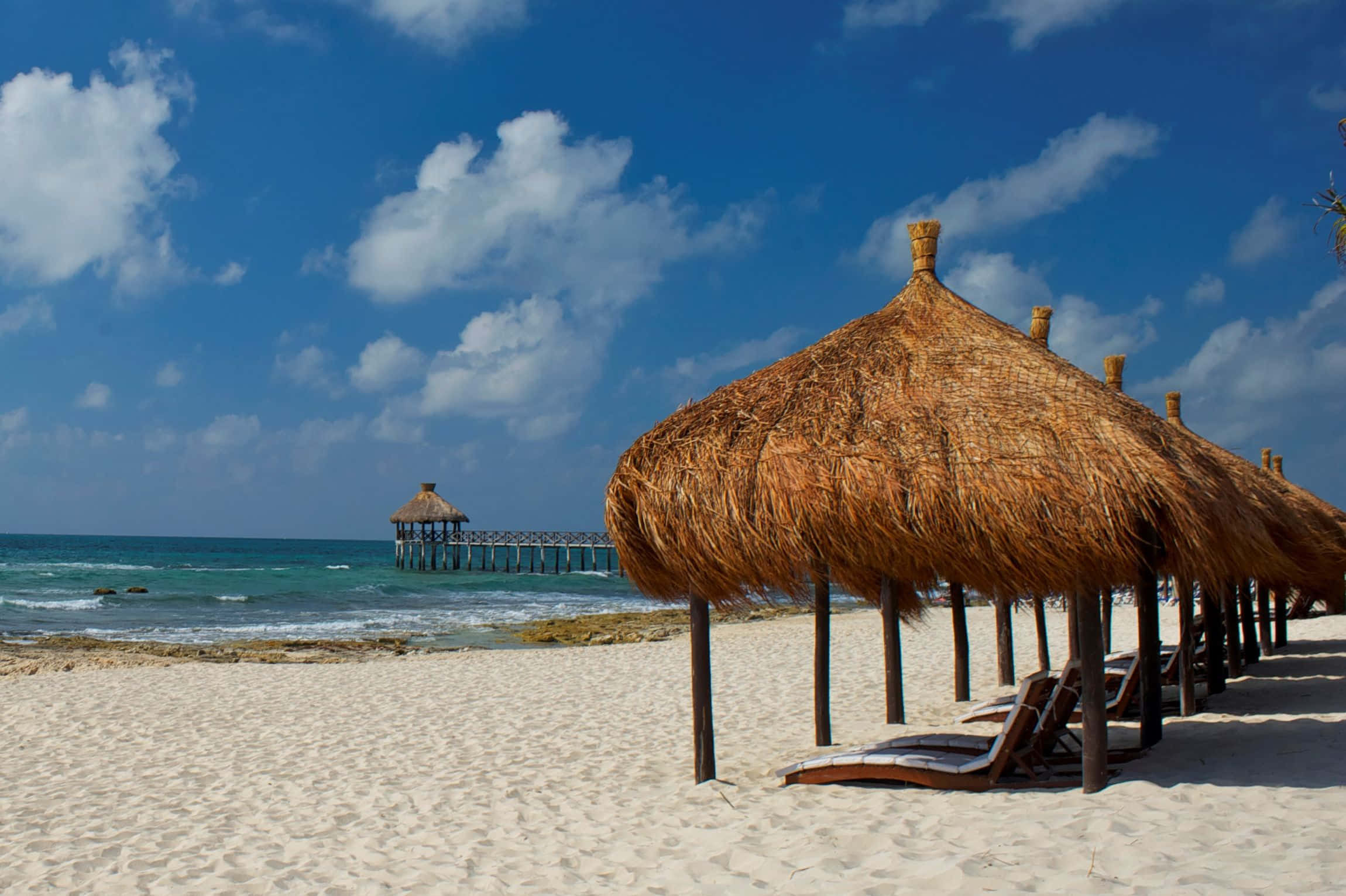 Mexican Beach 桌布 壁紙 — 茅草屋頂的小屋排列在純淨的白色沙灘上，天空湛藍，雲朵點綴，碧綠的海浪拍打著海岸。
