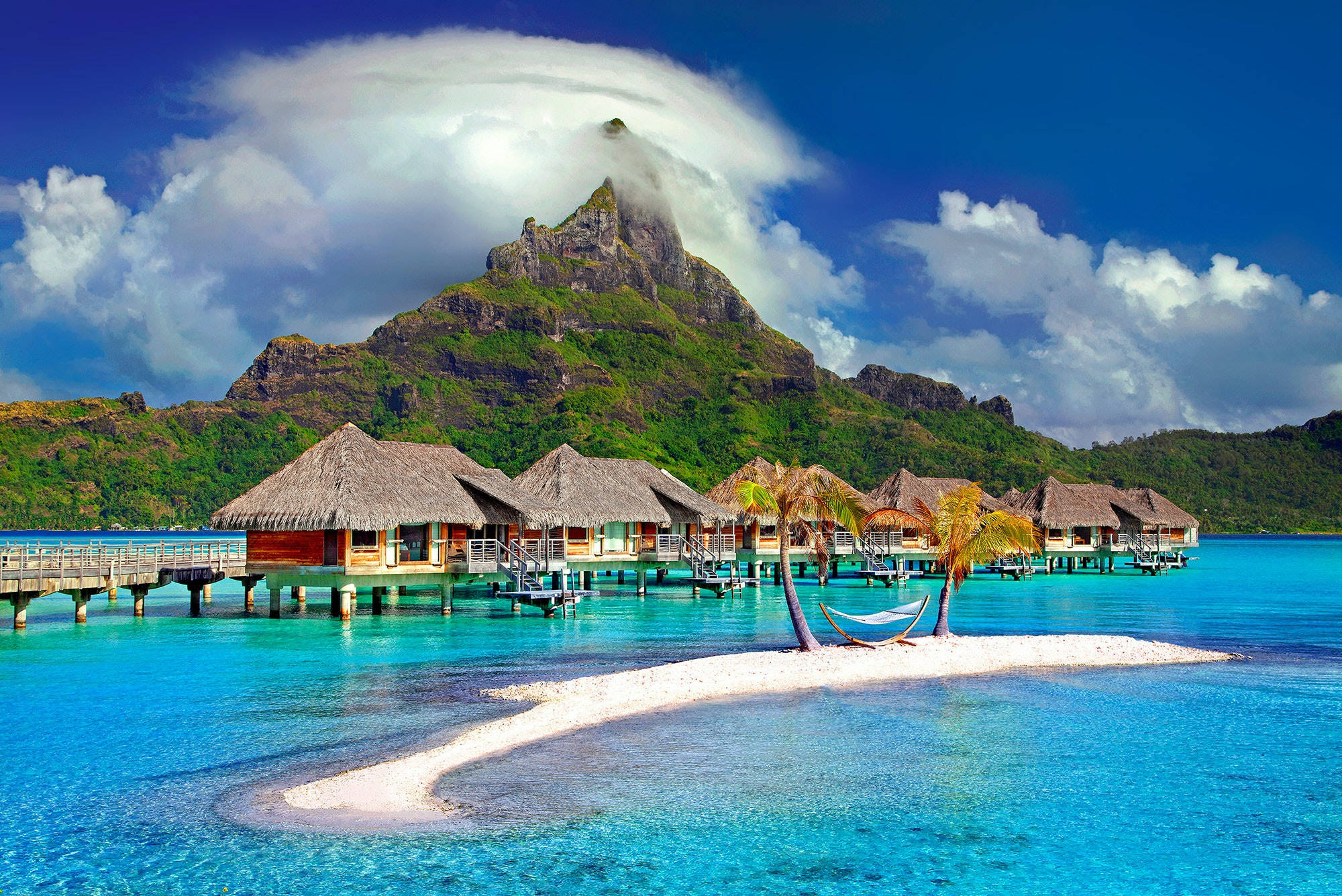 Beach Desktop 桌布 壁紙 — 茅草屋頂的水上平房漂浮在碧綠色的潟湖水面上，旁邊是白色沙洲，棕櫚樹下掛著吊床，背景是鬱鬱蔥蔥的綠色山脈，部分被雲霧籠罩，天空湛藍明亮。