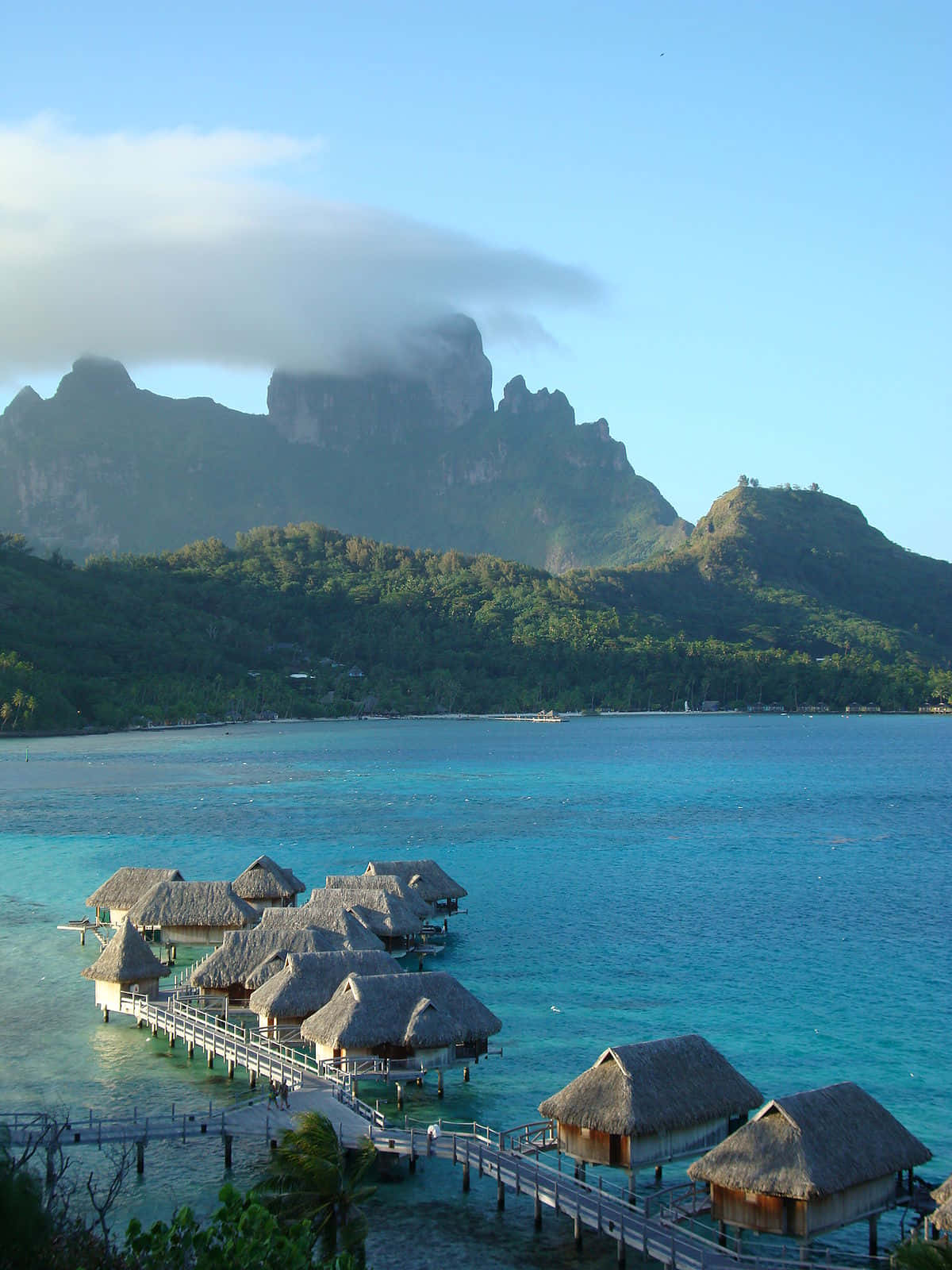 Bora Bora Beach 桌布 壁紙 — 茅草屋頂的水上平房漂浮在碧綠色的潟湖水面上，背景是部分被霧雲籠罩的茂密青山。