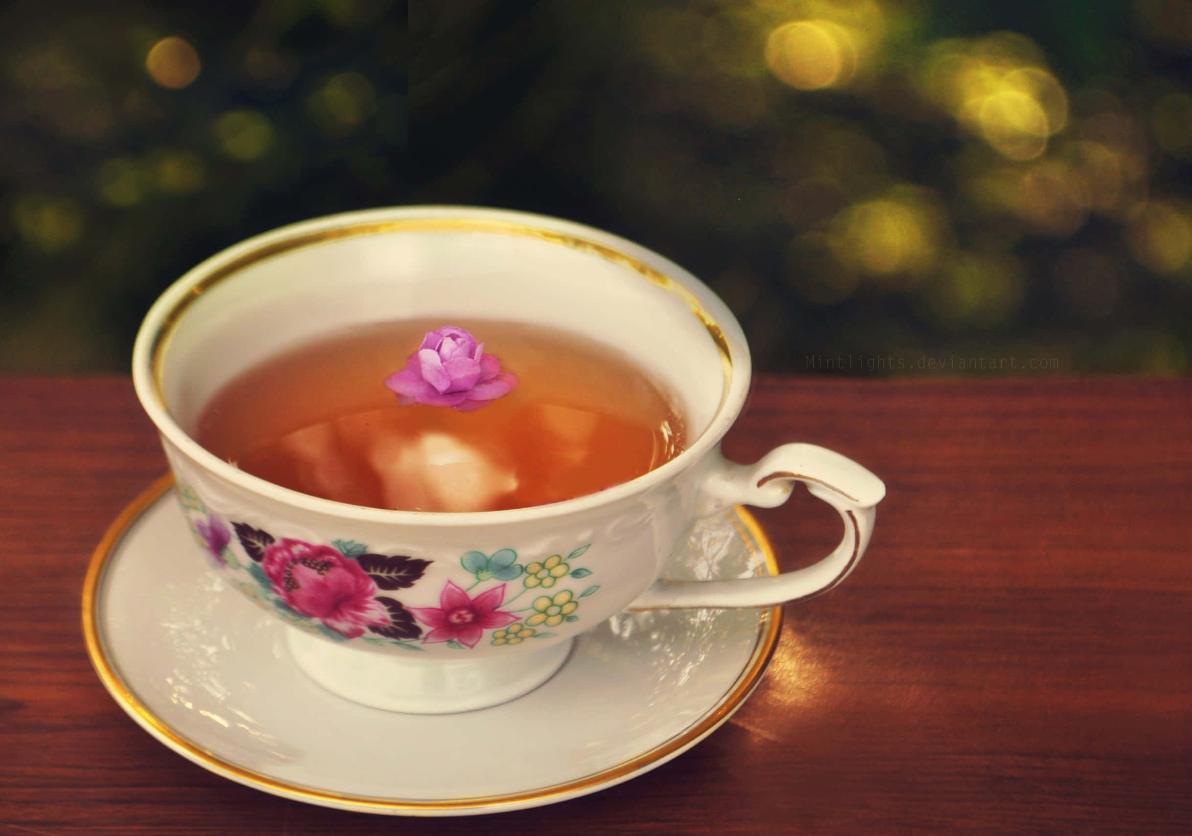 茶杯與柑橘花圖 壁紙 — 精緻的花卉茶杯，金色邊緣，盛載著琥珀色茶和一朵漂浮的粉紅玫瑰，背景是模糊的綠色散景。