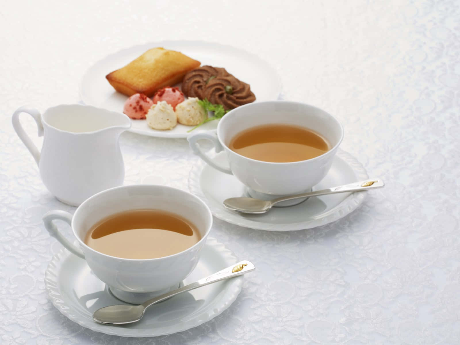 茶杯與茶具圖 壁紙 — 兩個白色茶杯，內有茶和匙羹，放在一個蕾絲枱布上，旁邊有一碟糕點。