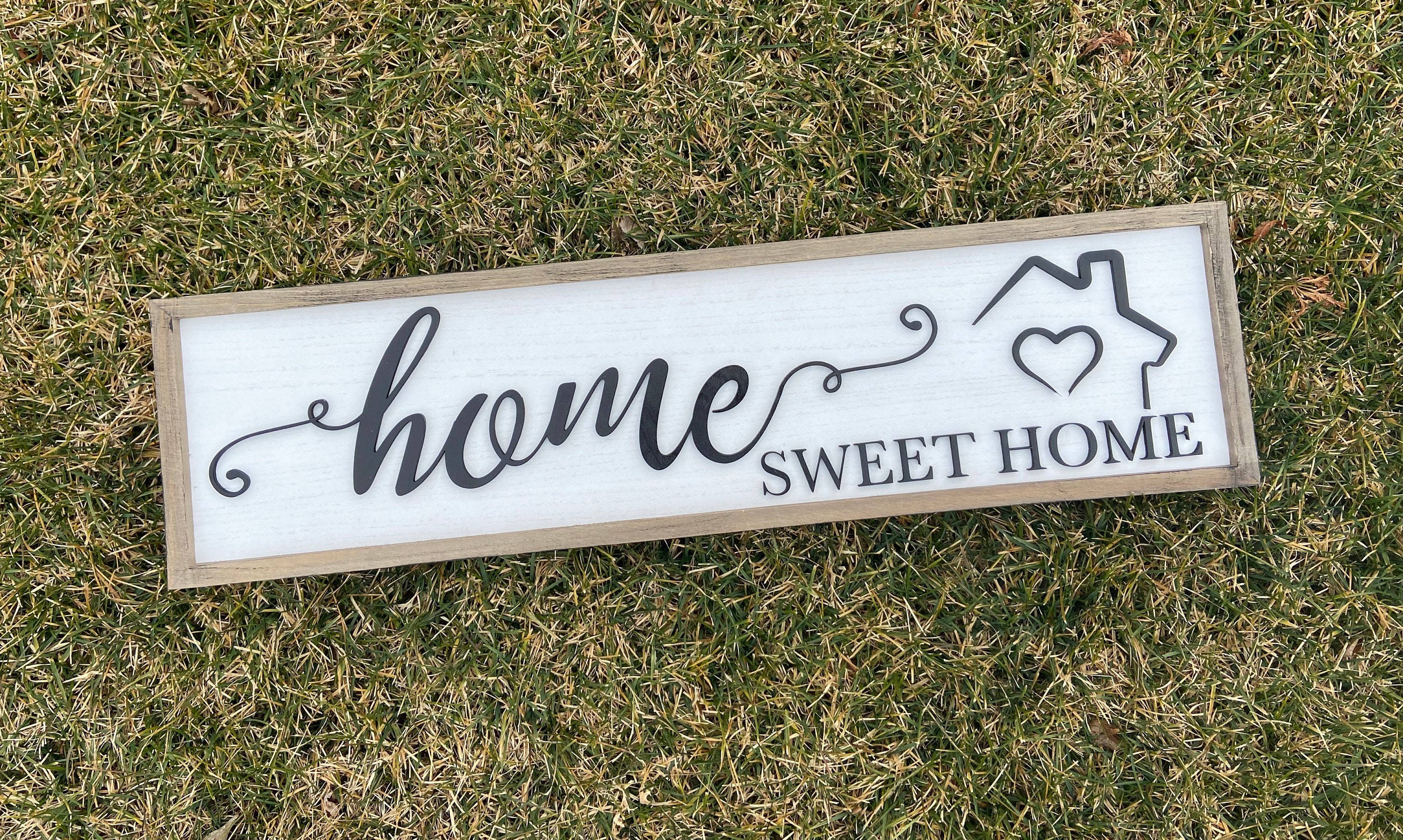 草地上的「家是甜蜜的家」標誌 壁紙 — 一塊寫有「home sweet home」的質樸木牌，帶有心形房屋圖標，放置在綠色和棕色的草地上。