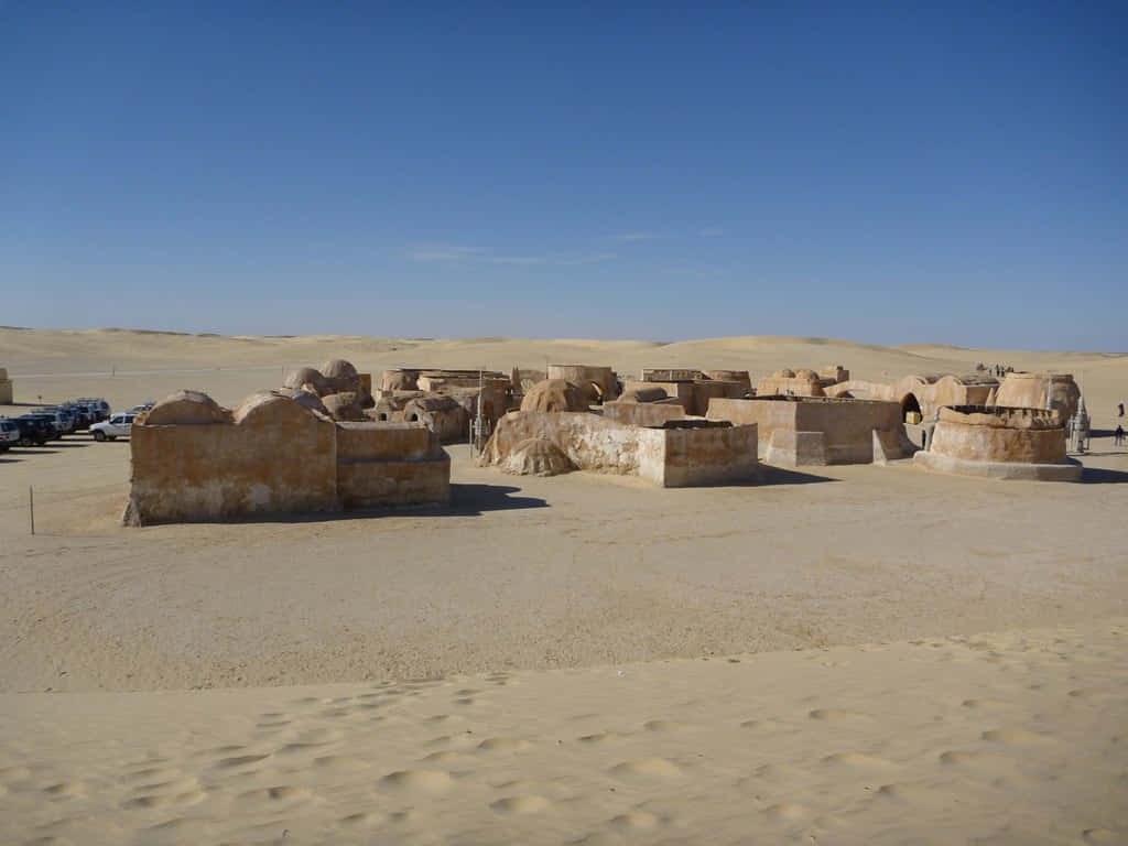 Mos Eisley 桌布 壁紙 — 荒涼的沙漠景觀，以莫斯艾斯利的標誌性圓頂建築為特色，在晴朗的藍天下，附近停有汽車。