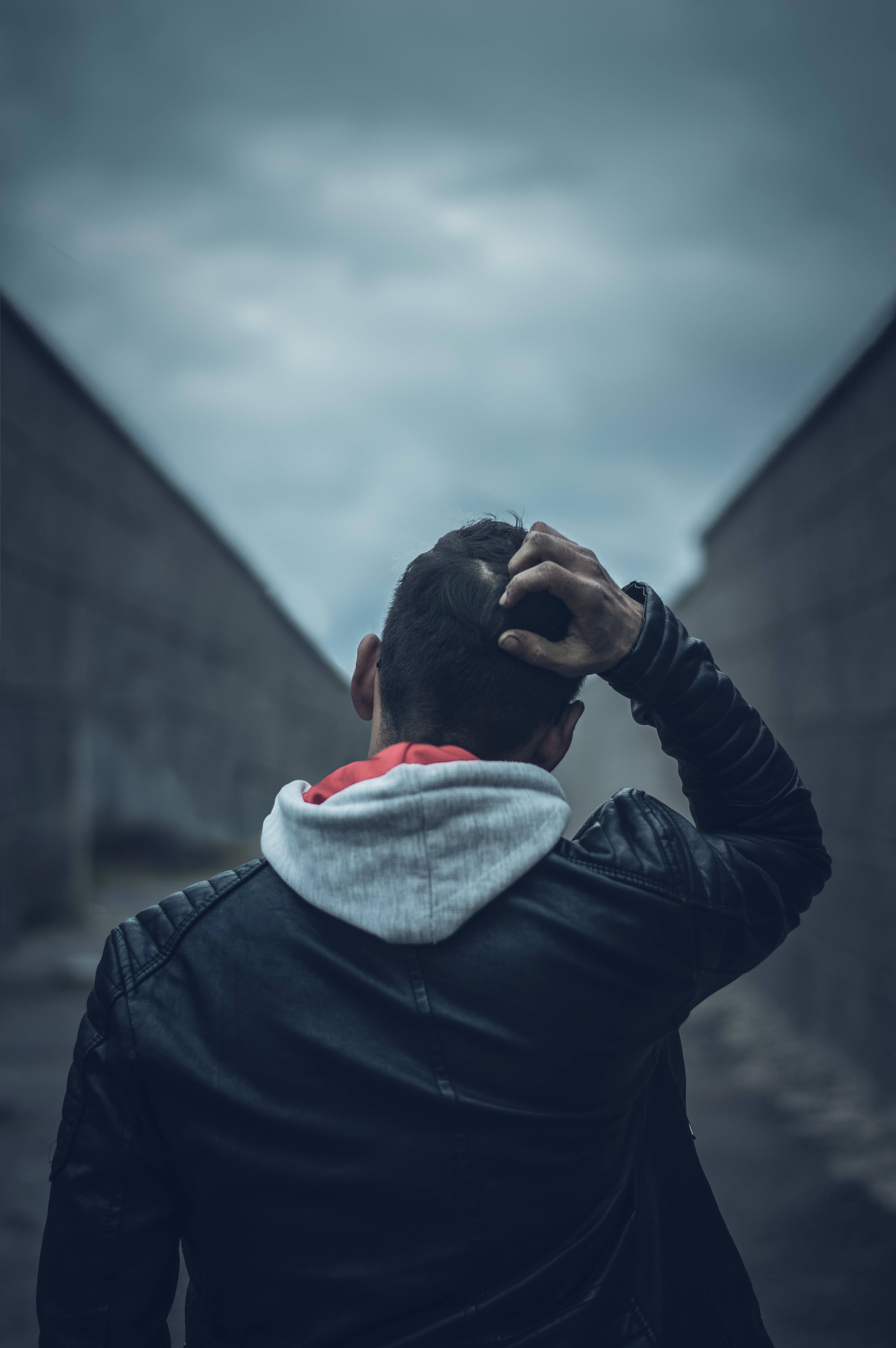 荒蕪的情感——一個孤寂少年對抗廣闊視野 壁紙 — 一個穿皮褸的男人背對鏡頭，手放在頭上，站在灰色天空下狹窄的混凝土小巷中。