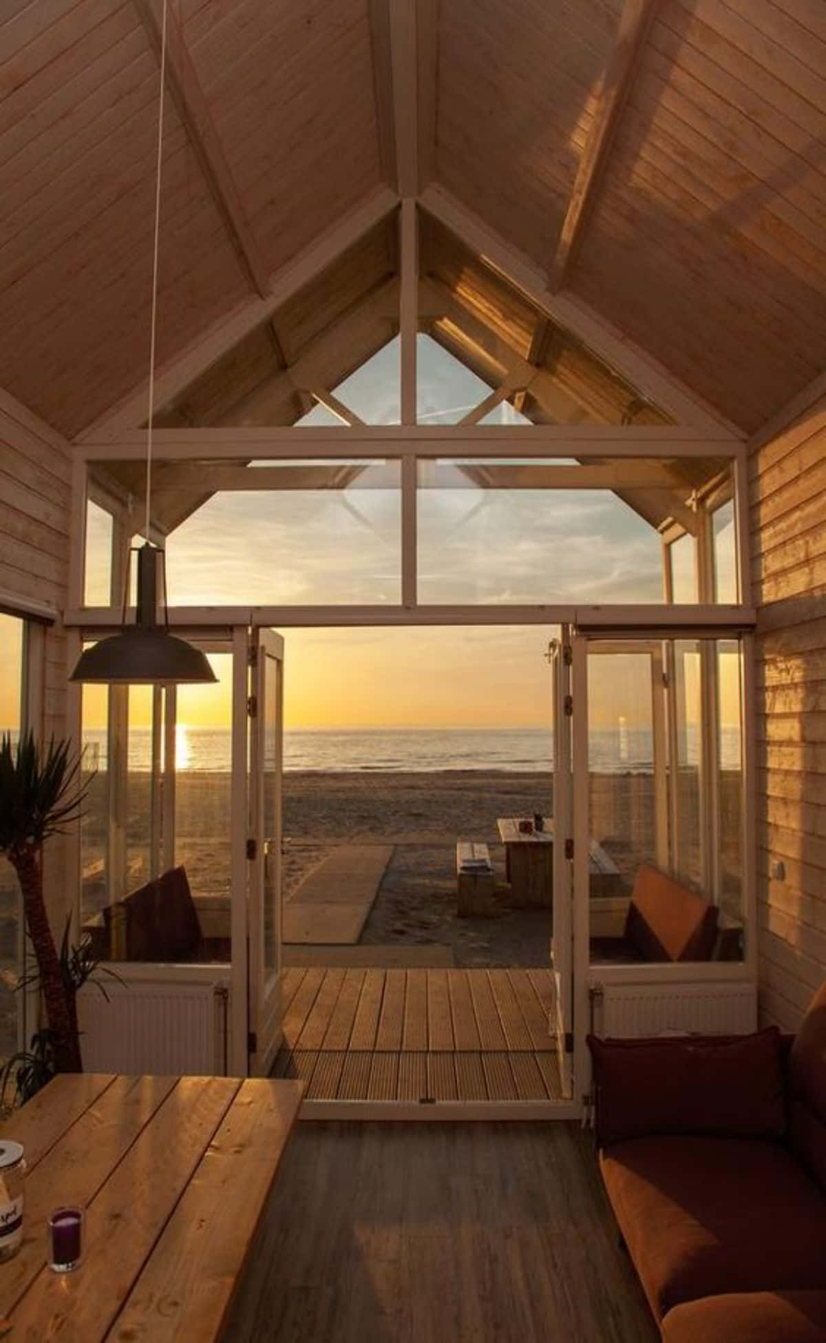 荷蘭鄉村小屋風景攝影圖 壁紙 — 溫暖的木質小屋內部通向寧靜的海灘，金色夕陽柔和光線透過玻璃門灑入。