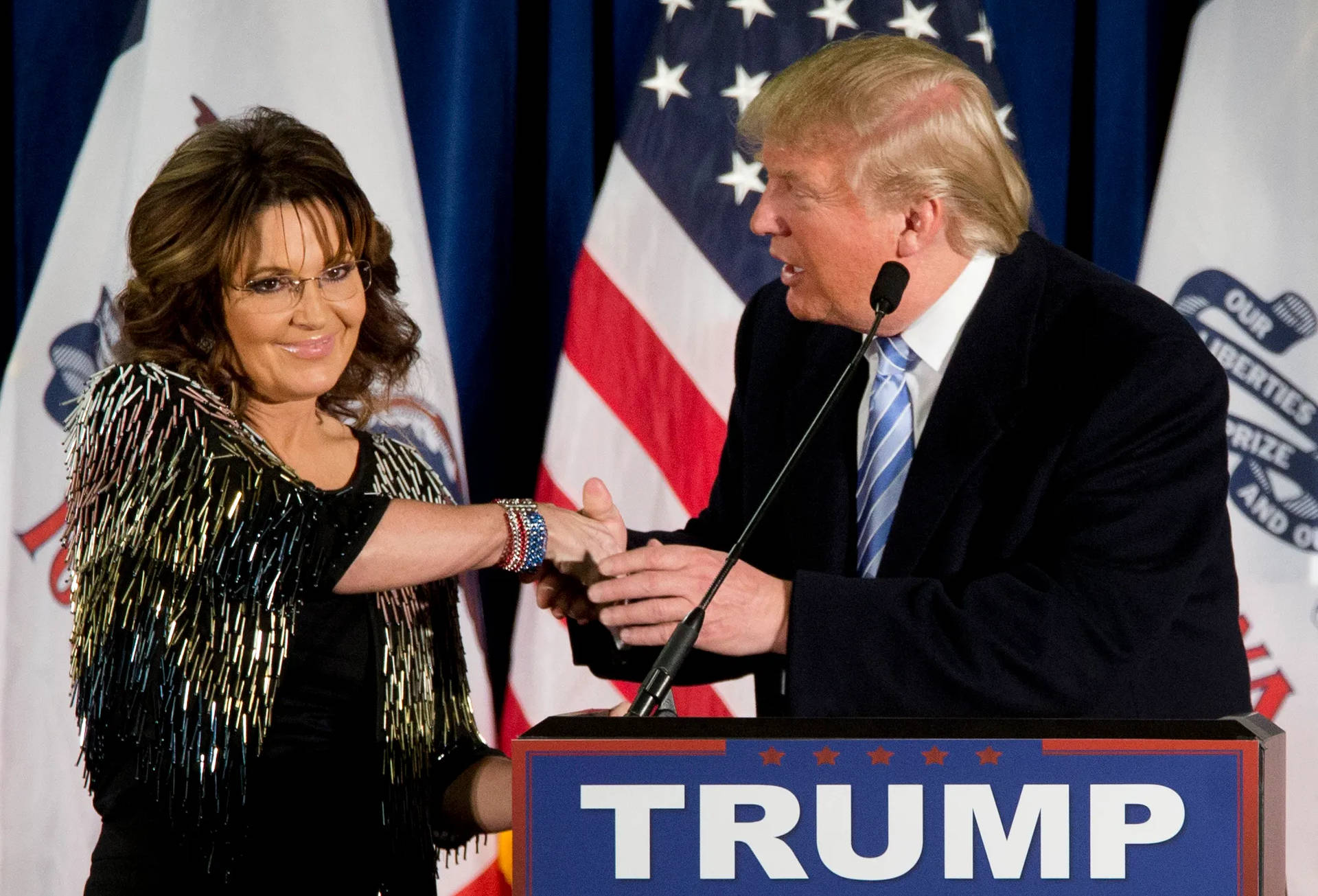Sara Lee 和 Donald Trump 在政治集會中握手，背景有旗幟和 Trump 講台。