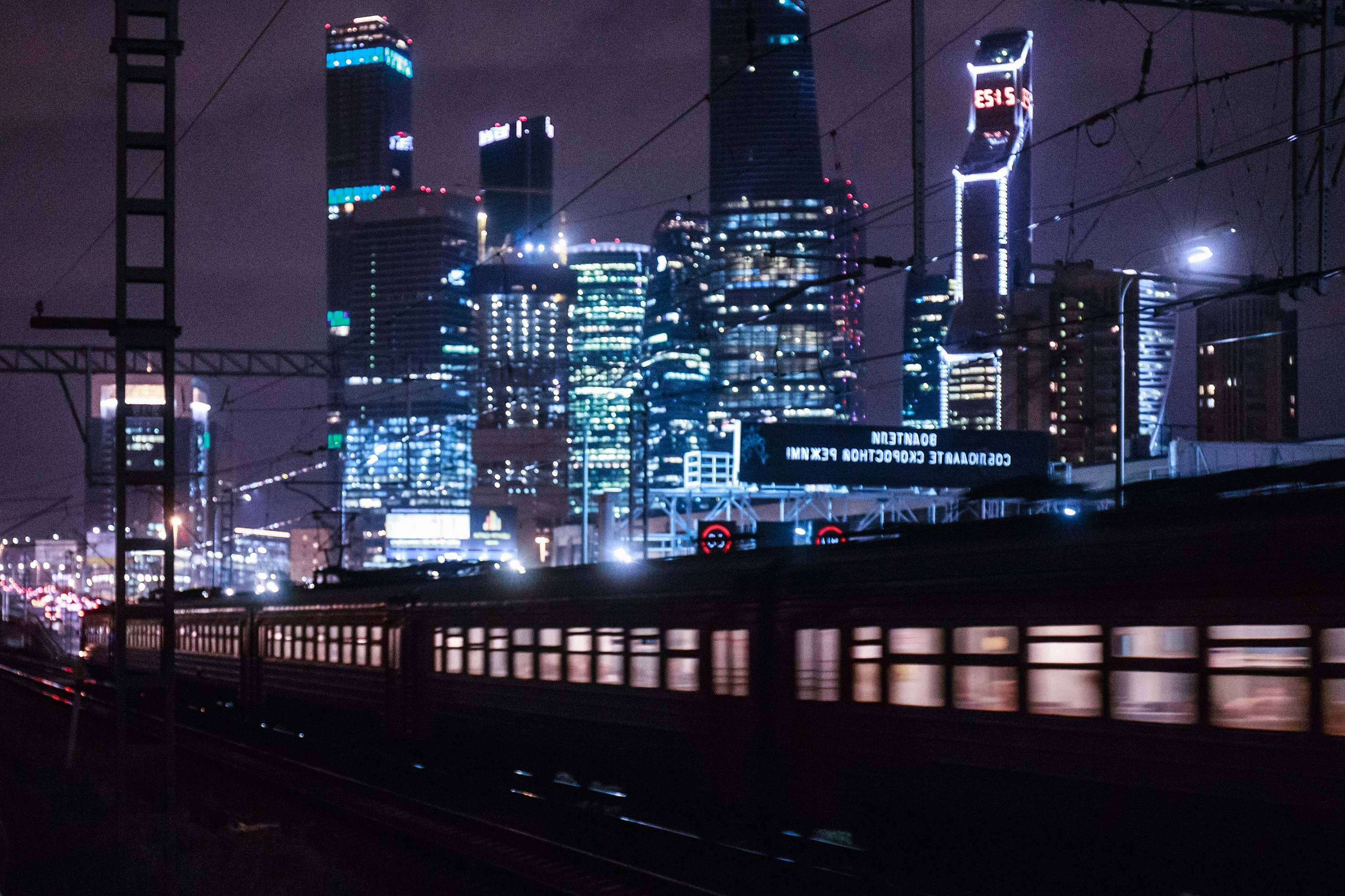 莫斯科俄羅斯都市列車美學 壁紙 — 一列高速行駛的火車在夜晚霓虹燈閃爍的高聳城市天際線下模糊掠過。