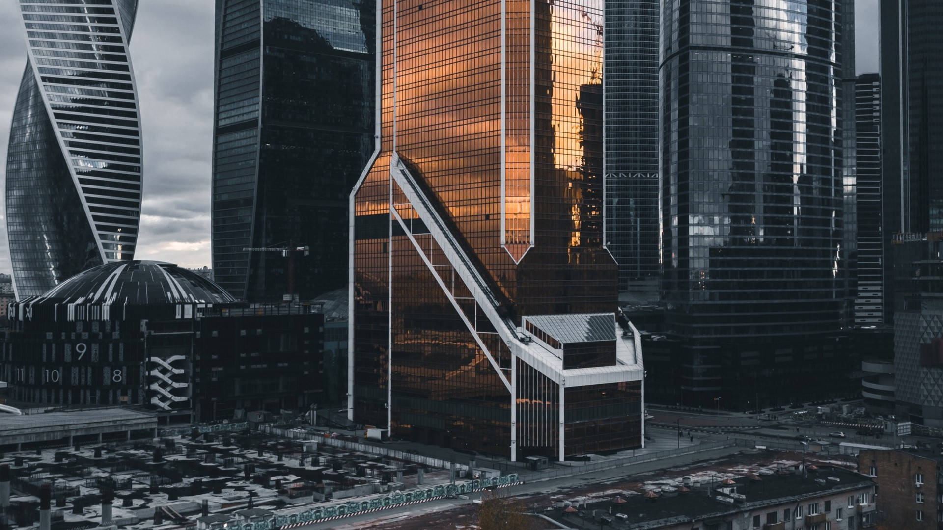 莫斯科水銀塔銅色外觀 壁紙 — 現代玻璃摩天大樓在密集的城市天際線中，映照出金色夕陽色調，背景是陰沉的灰色天空。