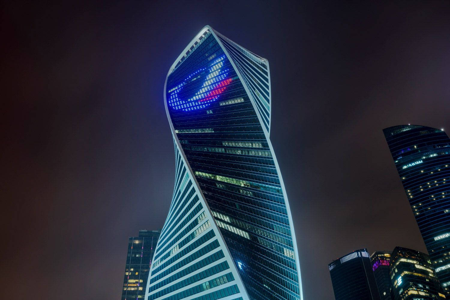 莫斯科演化塔夜景 壁紙 — 一座高聳扭曲的摩天大樓在現代城市景觀中，以藍色和紅色的燈光在黑暗的夜空中閃耀。