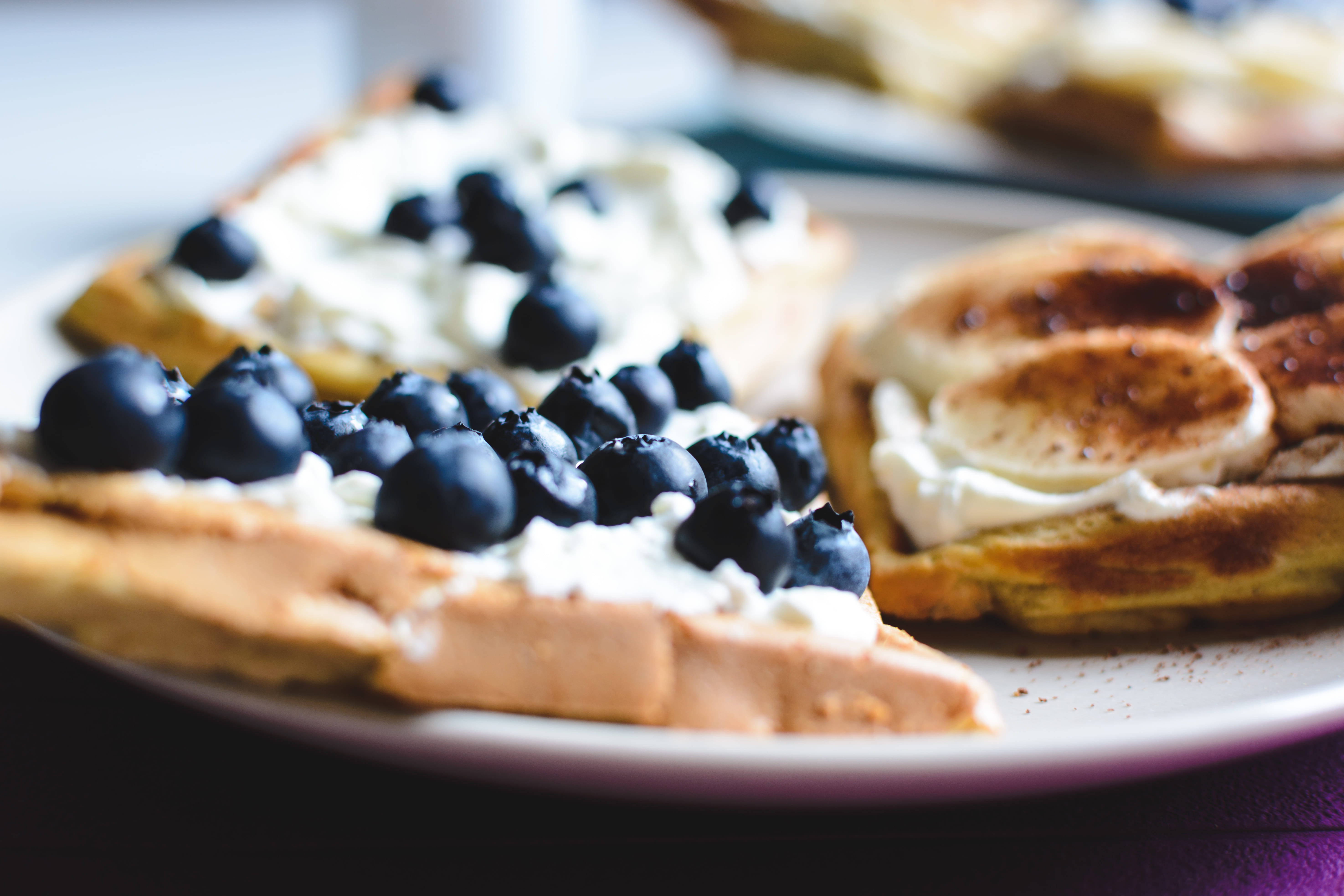 華夫餅搭配藍莓與香蕉 壁紙 — 金黃色的窩夫配上忌廉狀的鮮奶油和飽滿的藍莓，放在白色碟上，賣相誘人。