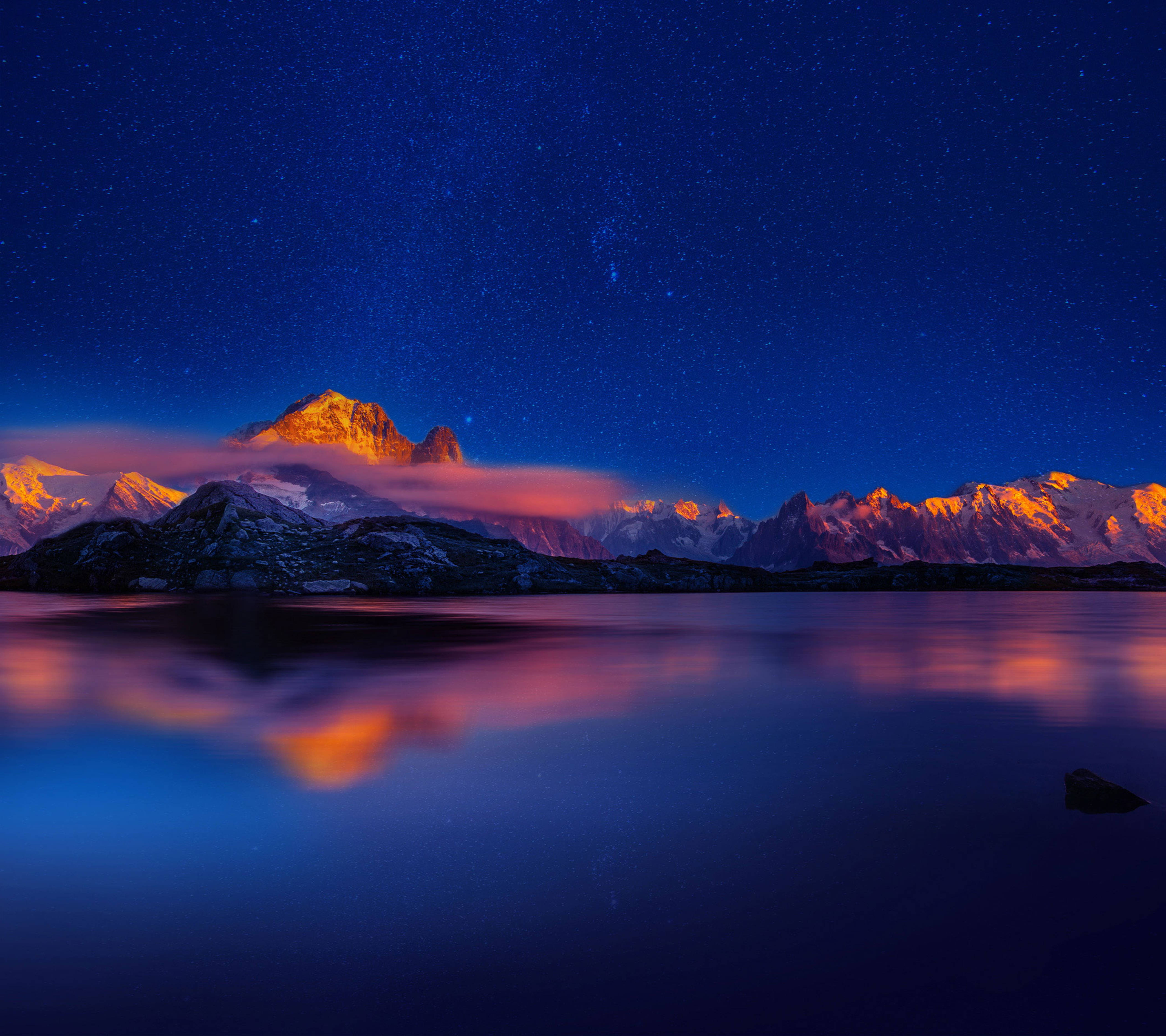 華為P9夜山 壁紙 — 星空之夜在積雪的山峰上閃耀，倒映在平靜的湖面上，伴隨著橙色的雲彩。