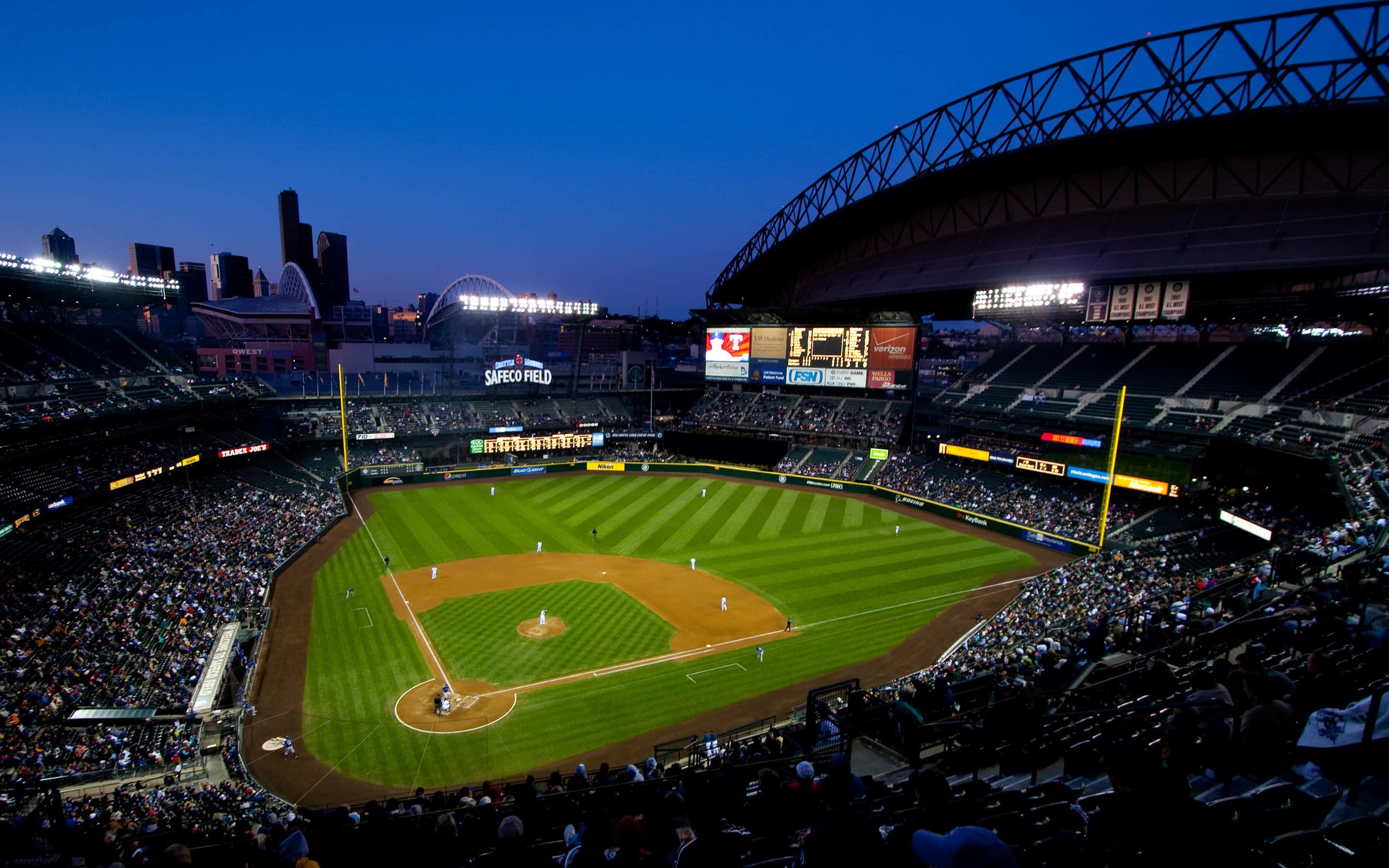 華盛頓 T-Mobile Park 棒球場景觀背景 壁紙 — 黃昏時的棒球場，燈光照亮的場地和擠滿觀眾的看台，背景是逐漸變暗的城市天際線。